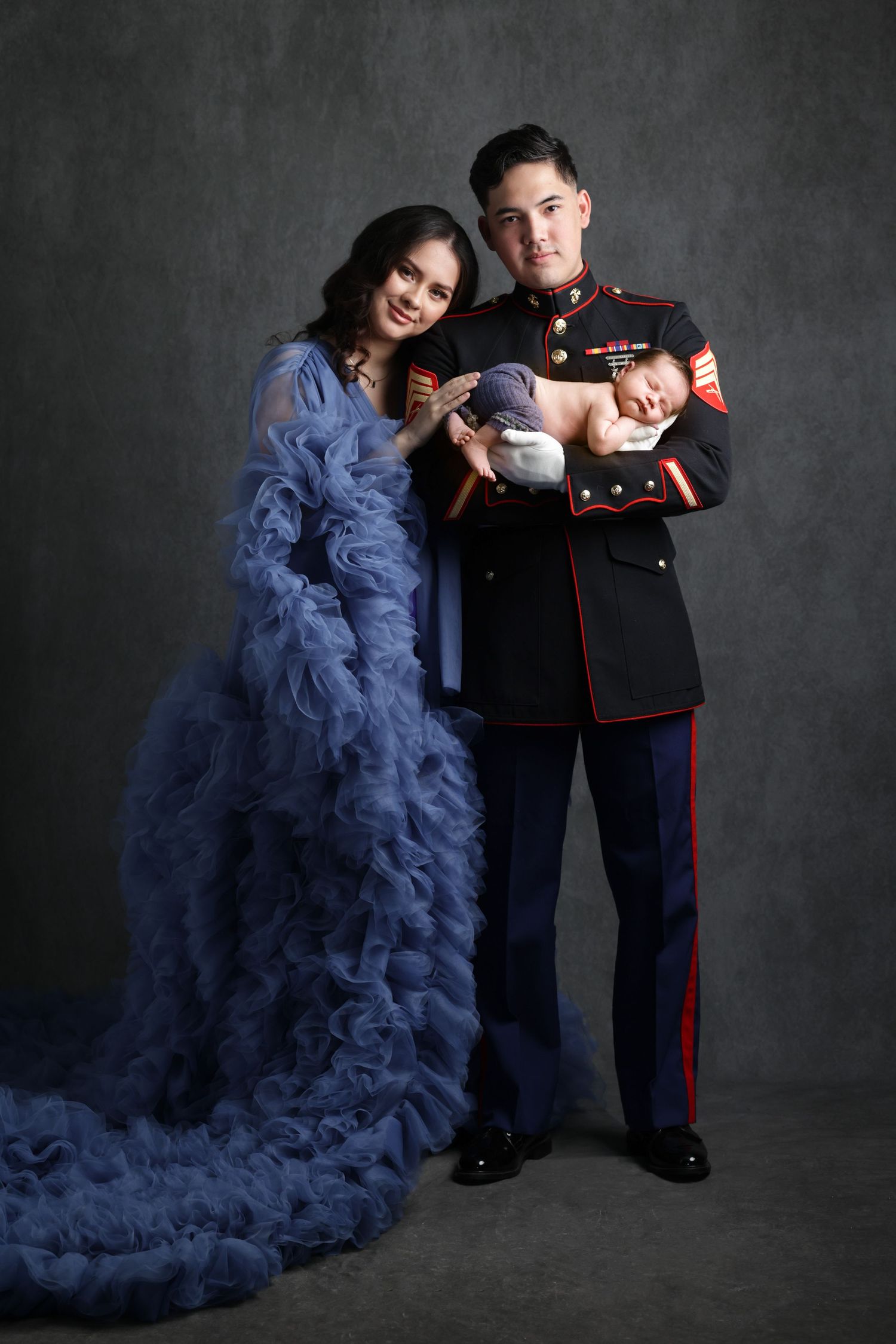 Marine in dress uniform holds newborn beside woman in blue ruffled gown in elegant studio portrait.