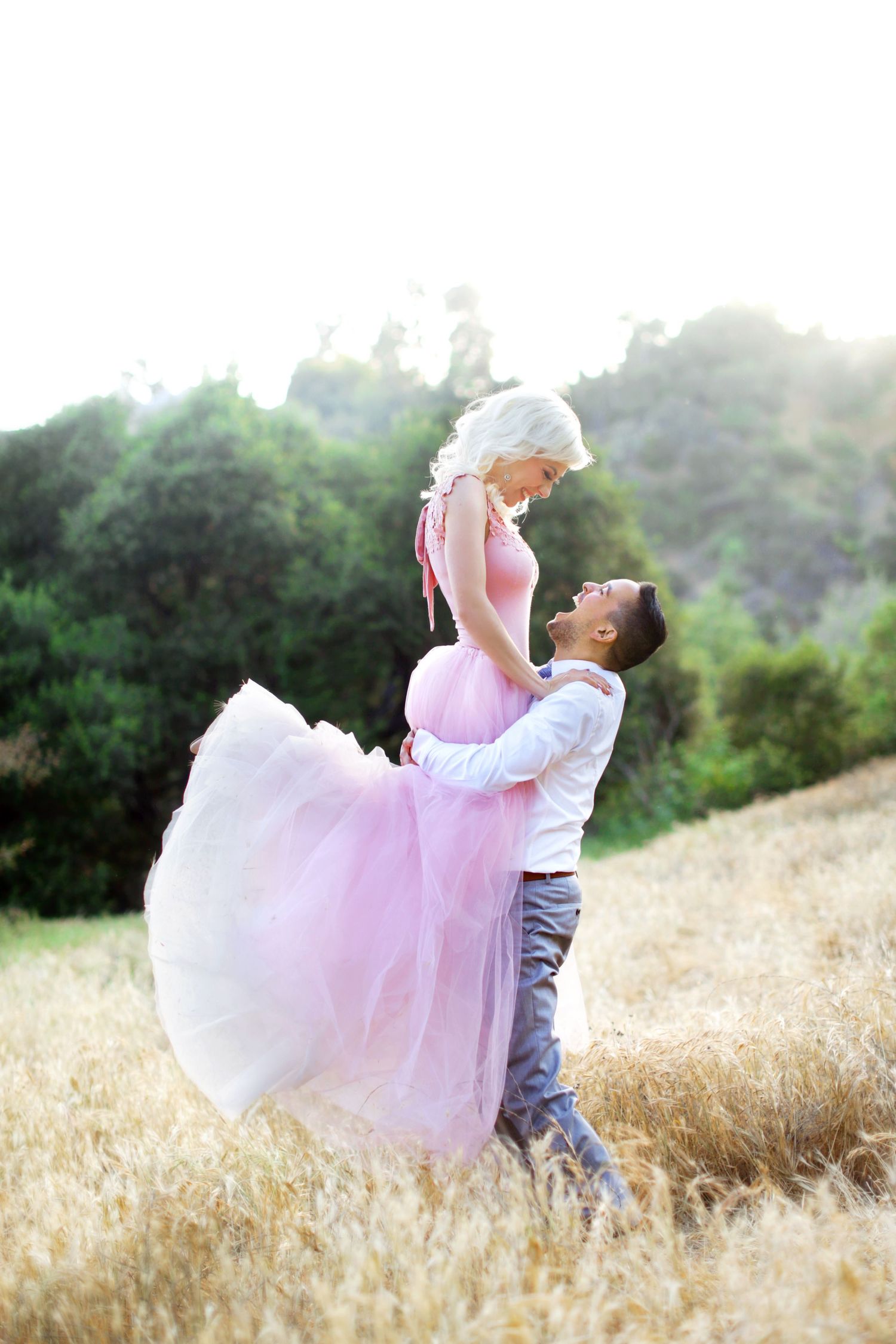 A man lifts a woman in a pink tulle dress in a golden field, surrounded by green trees in soft sunlight.