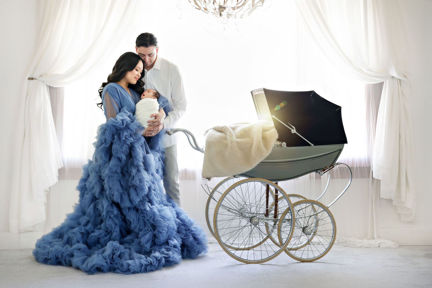 Couple in elegant attire with newborn baby beside a vintage pram in a bright white studio setting.