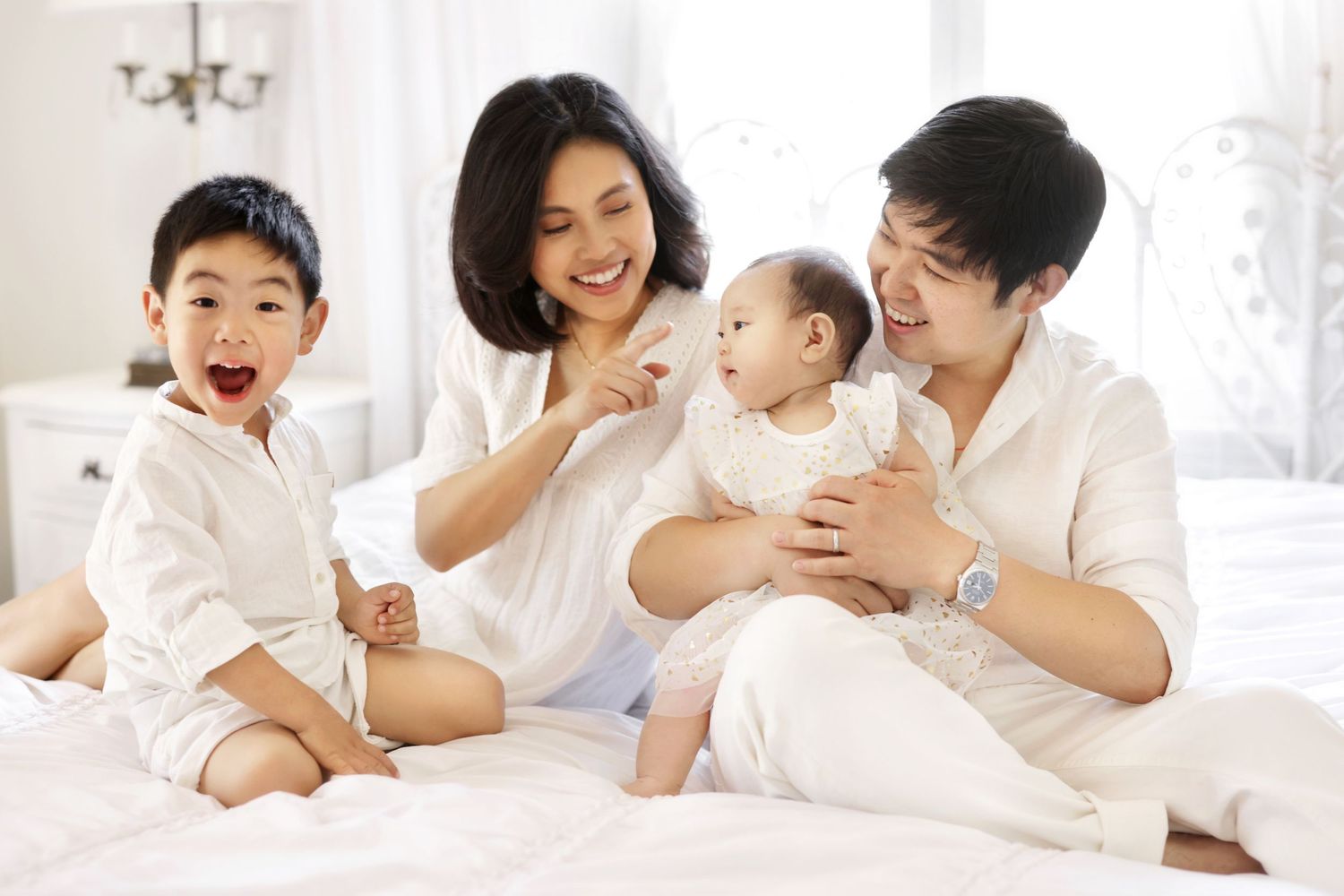 Happy Asian family with two children sitting on a white bed, parents smiling at their baby and toddler son.