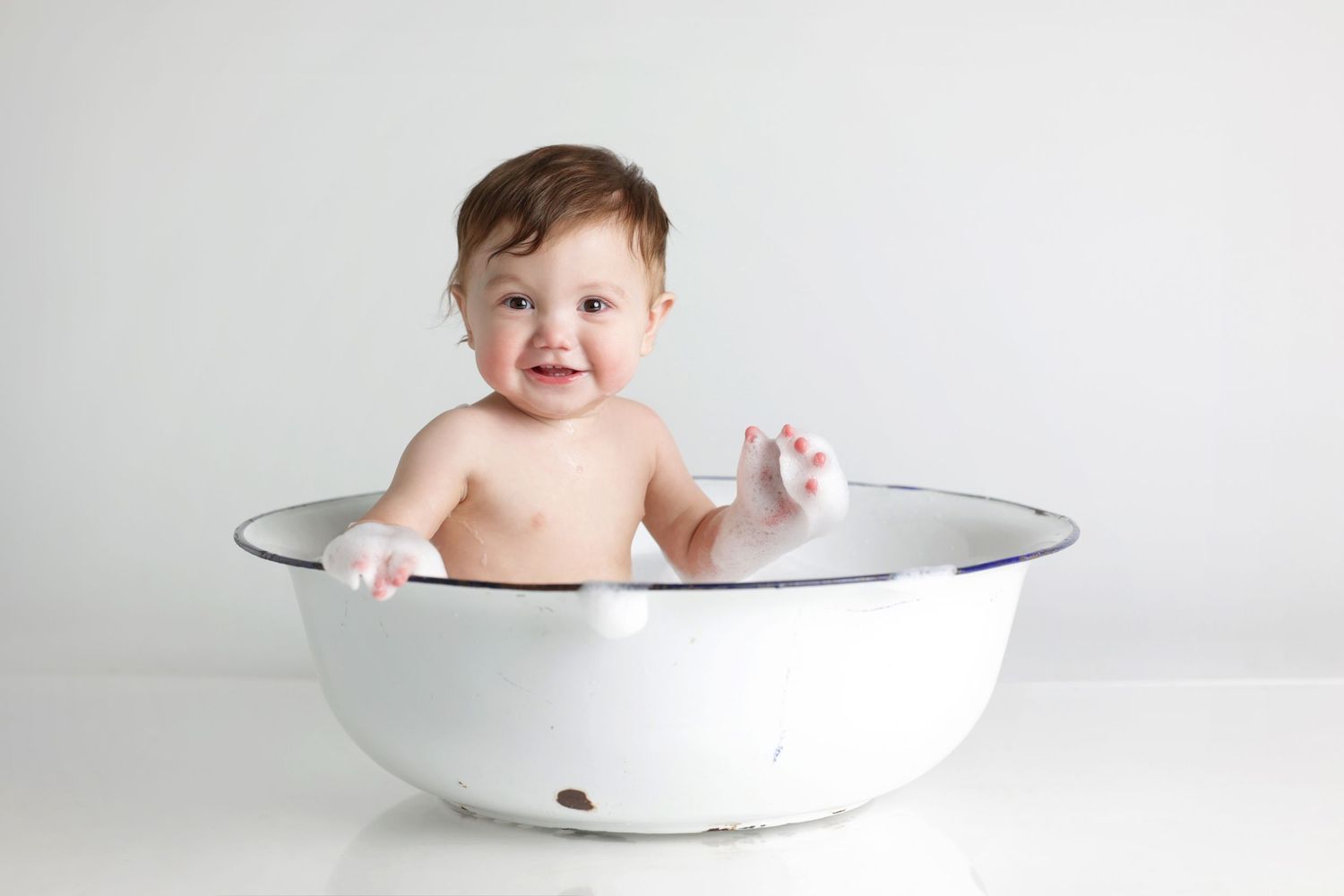 Smiling baby sitting in a white enamel basin tub, enjoying bath time against a clean white background.