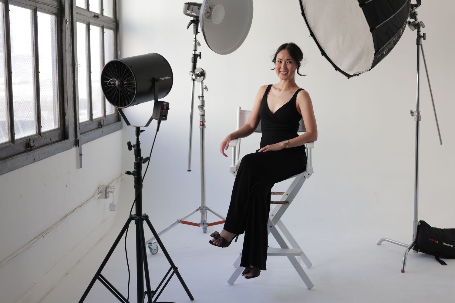 Woman in black outfit seated in director's chair surrounded by professional studio lighting equipment.