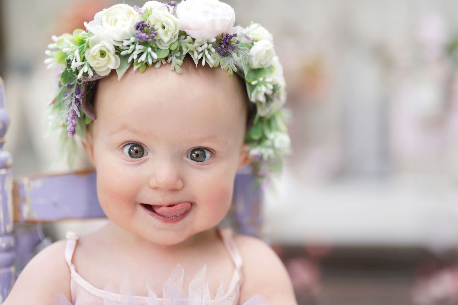 Smiling baby girl wearing a floral crown with white flowers and greenery, dressed in a pink outfit.