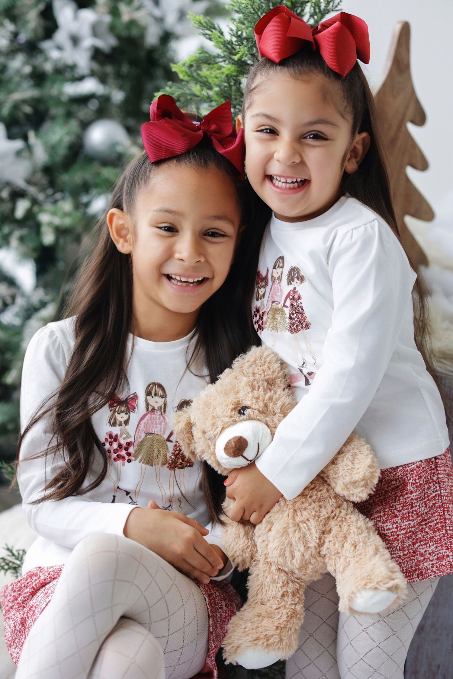 Two young girls in matching Christmas outfits with red bows smile while holding a teddy bear near a decorated tree.