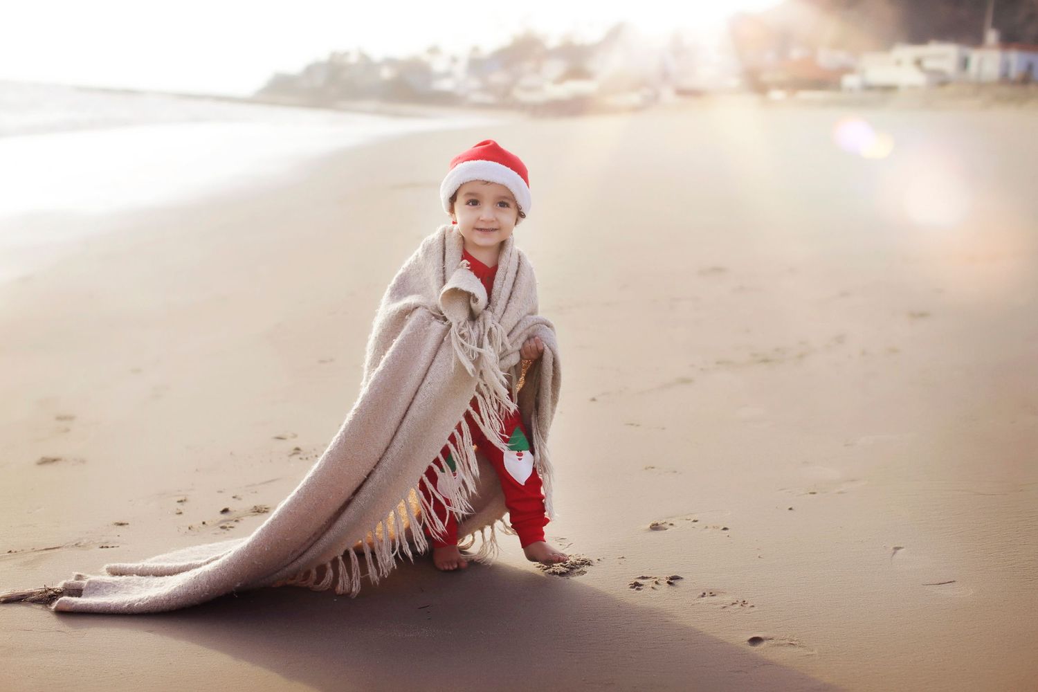 Child in Santa hat wrapped in blanket sits on sandy beach with sunlight glowing in background.