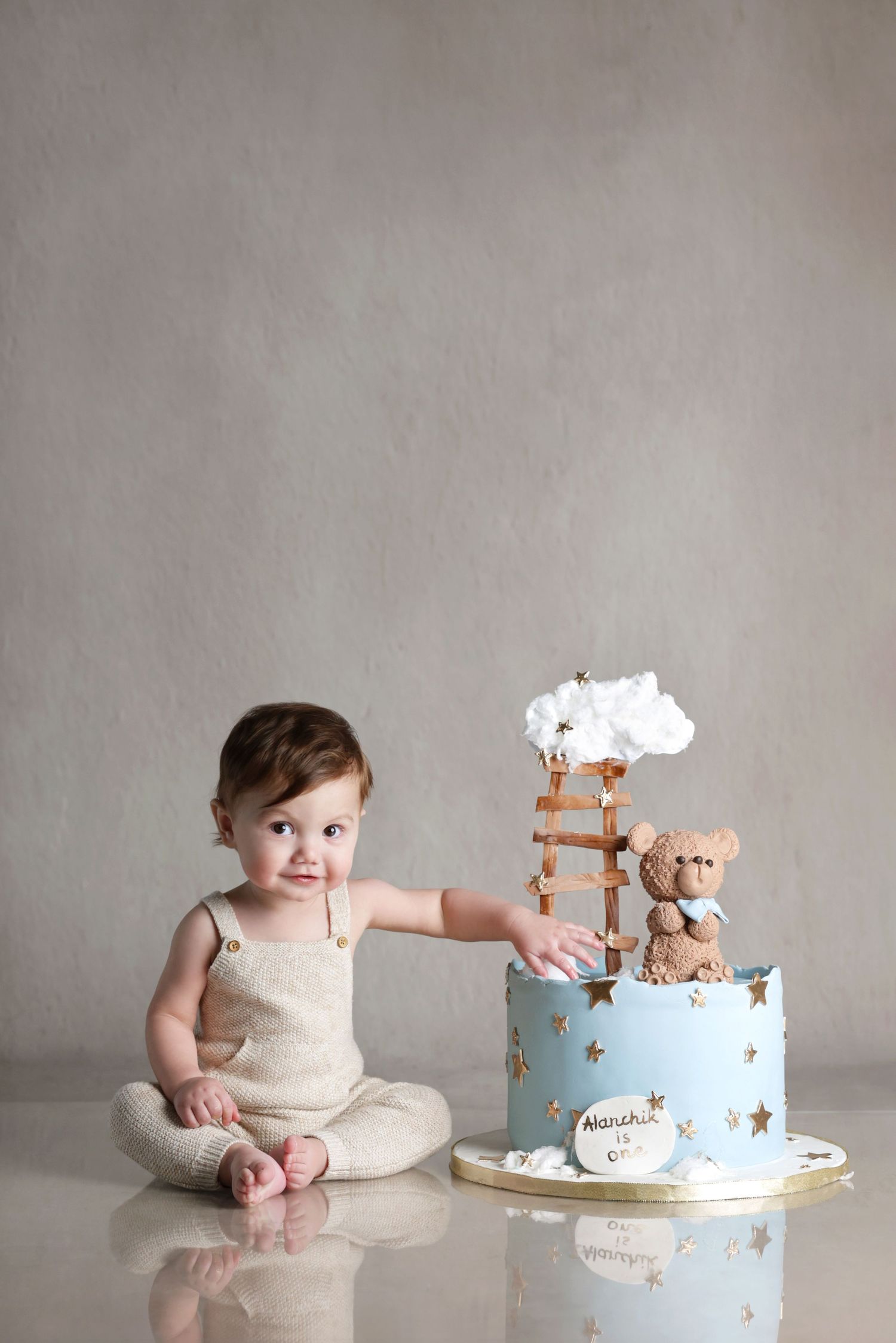 Cute baby in beige romper reaches toward blue first birthday cake with teddy bear topper.