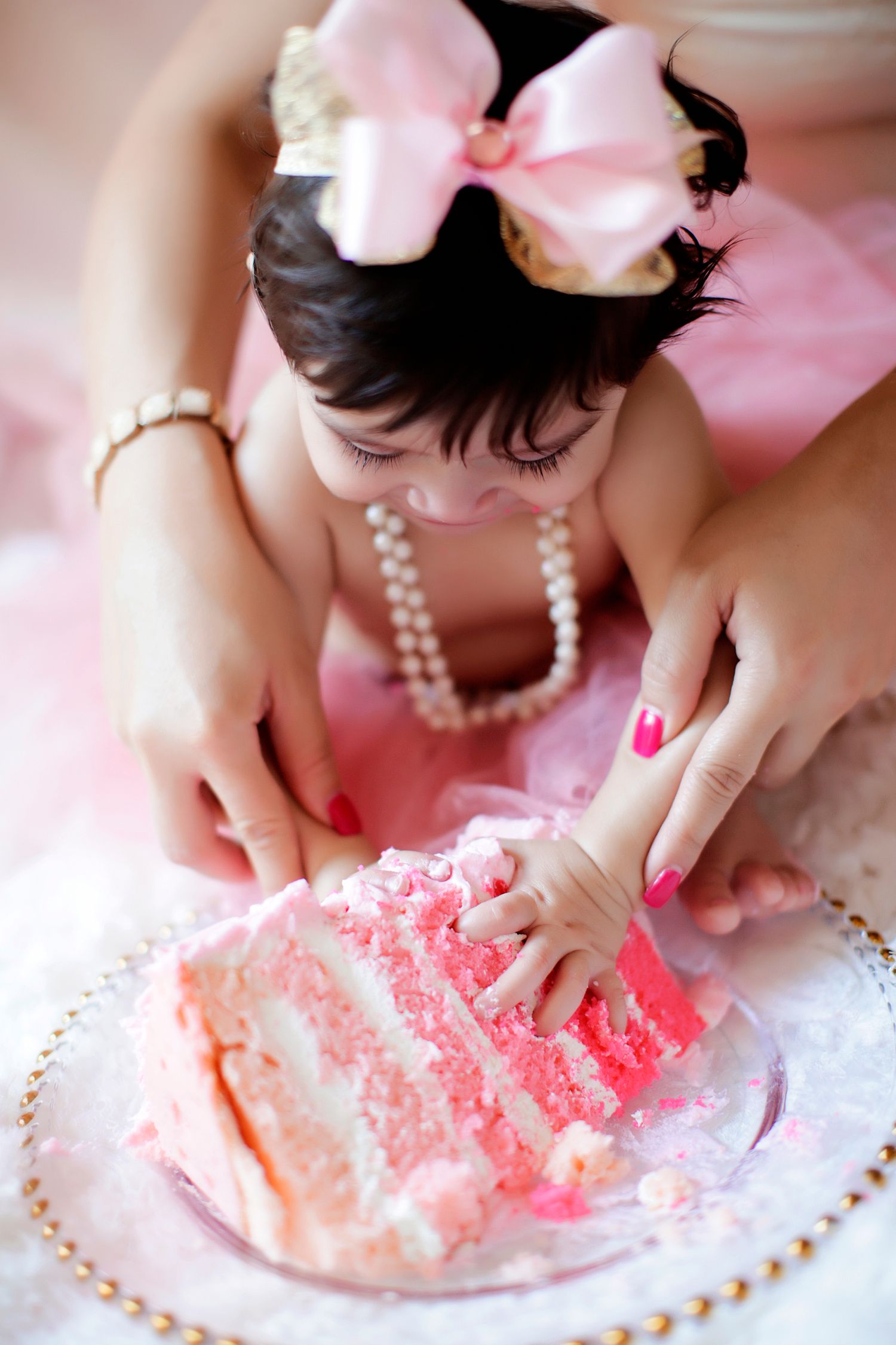 Baby girl in pink tutu and pearl necklace smashing her first birthday cake with adult hands guiding her.