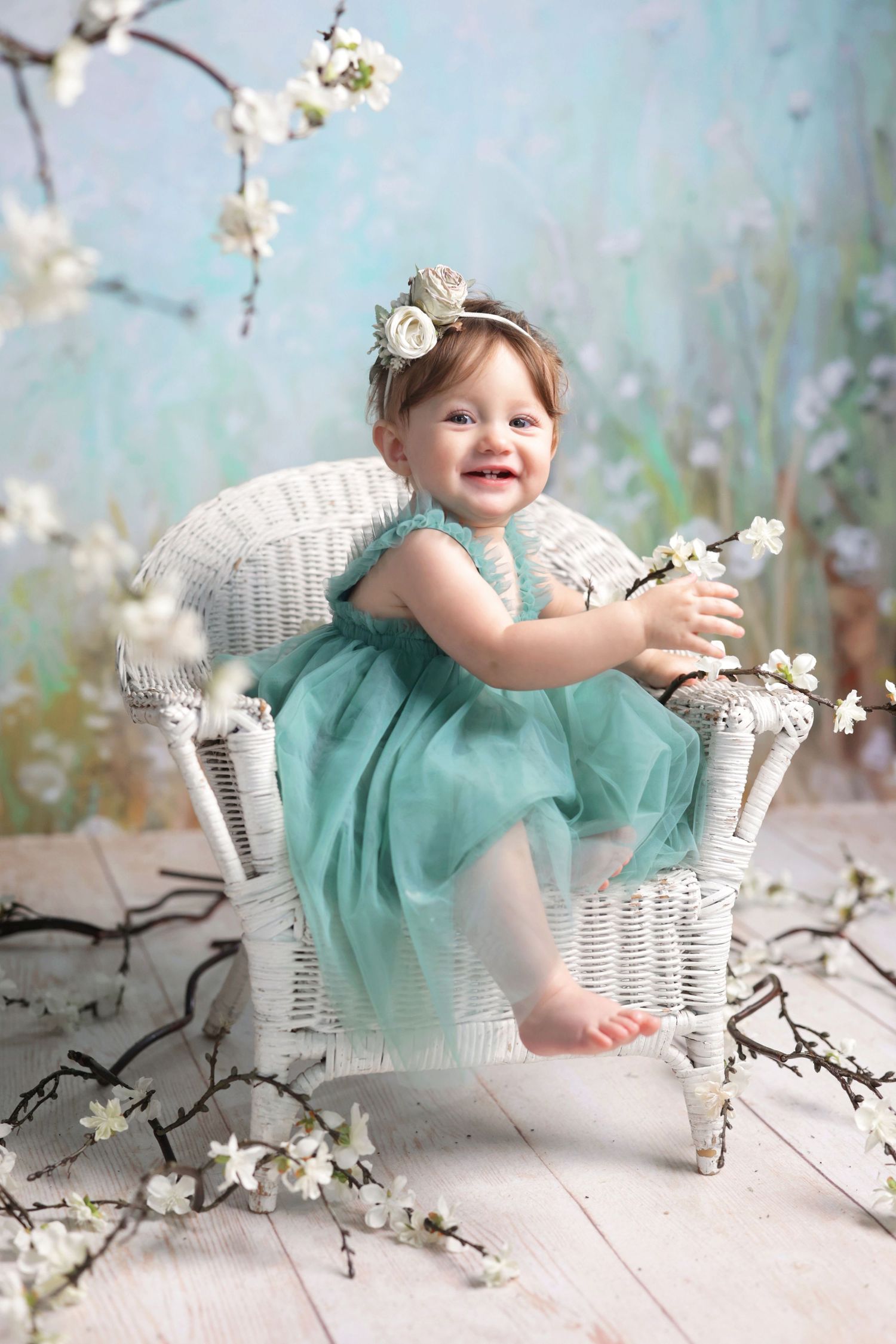 Smiling baby girl in teal tulle dress sitting in white wicker chair surrounded by white cherry blossom branches.