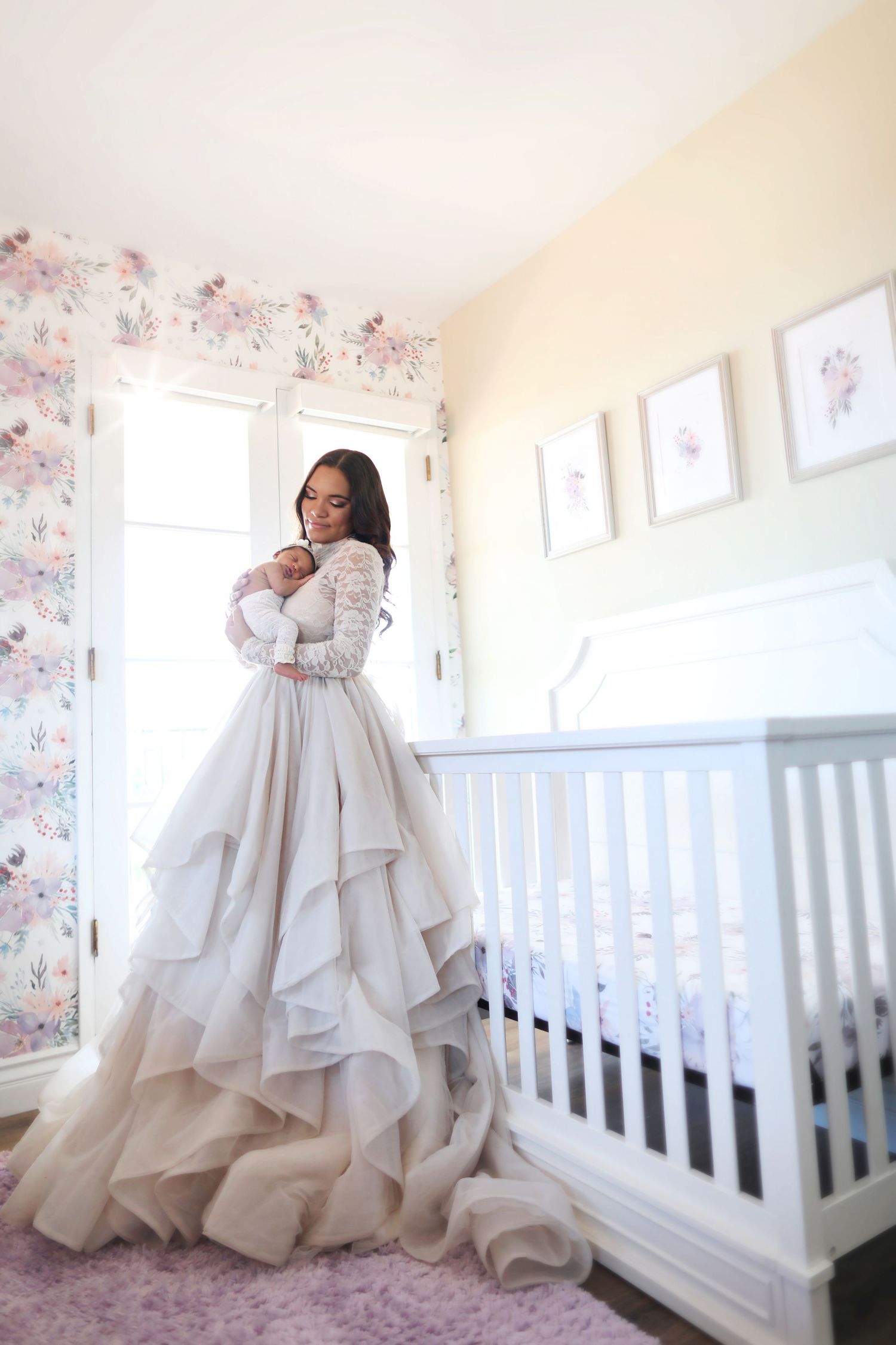 Woman in elegant layered bridal gown holding newborn baby beside white crib in floral nursery room.