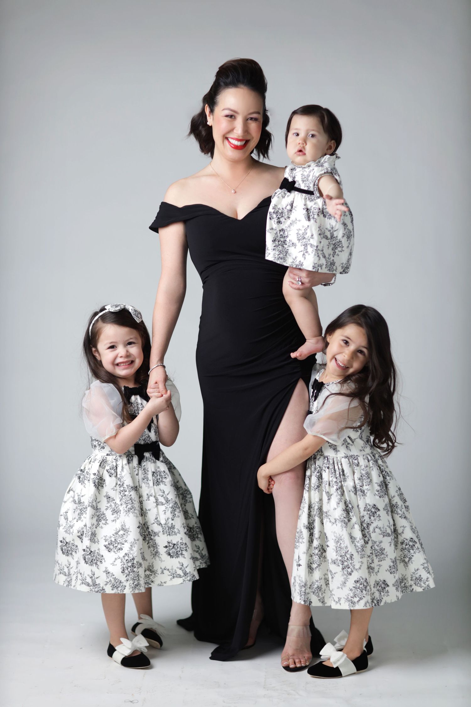 A woman in a black gown poses with three young girls in matching floral dresses in a studio portrait.