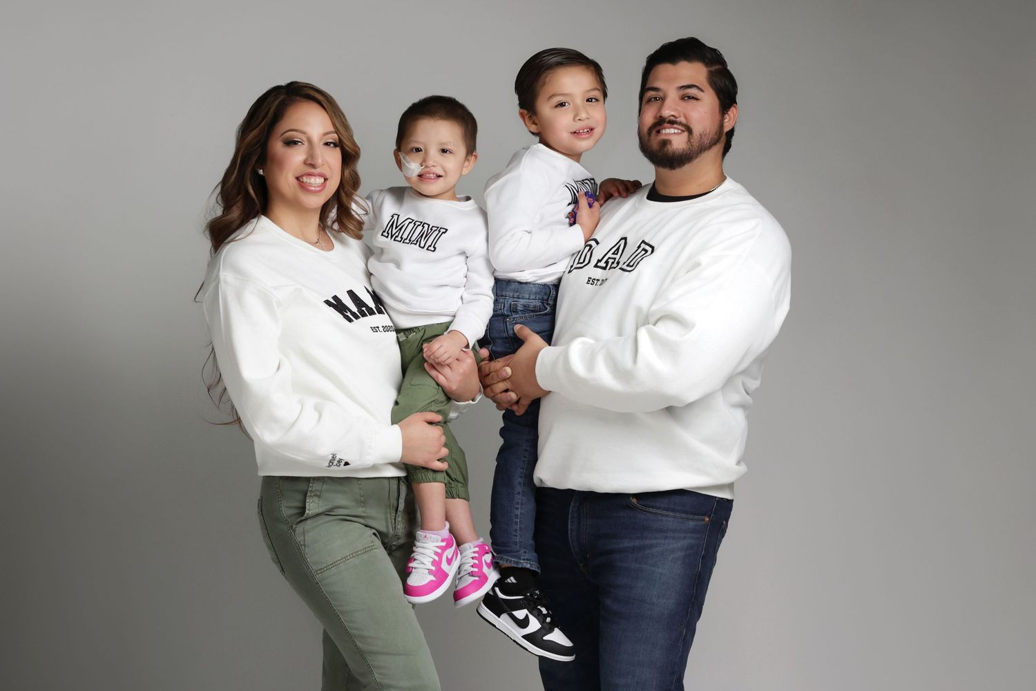 Family of four in matching white sweatshirts posing for a portrait, parents holding two young children.