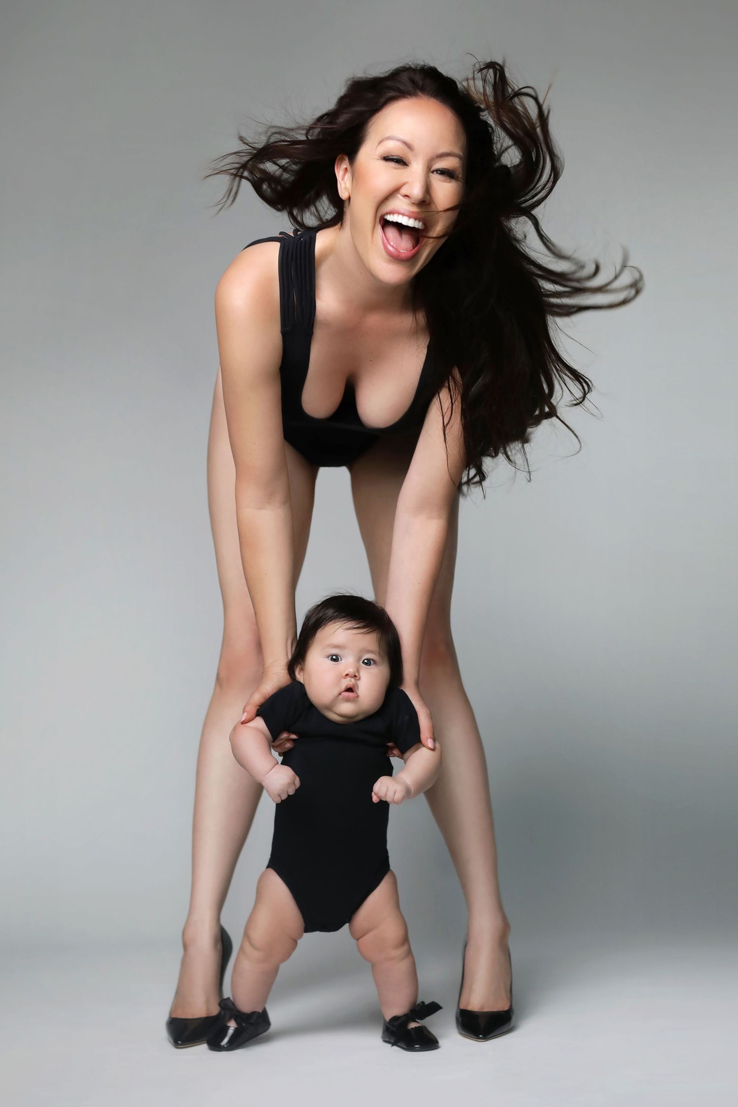 Laughing woman in black bodysuit helping baby stand in matching outfit on gray studio background.