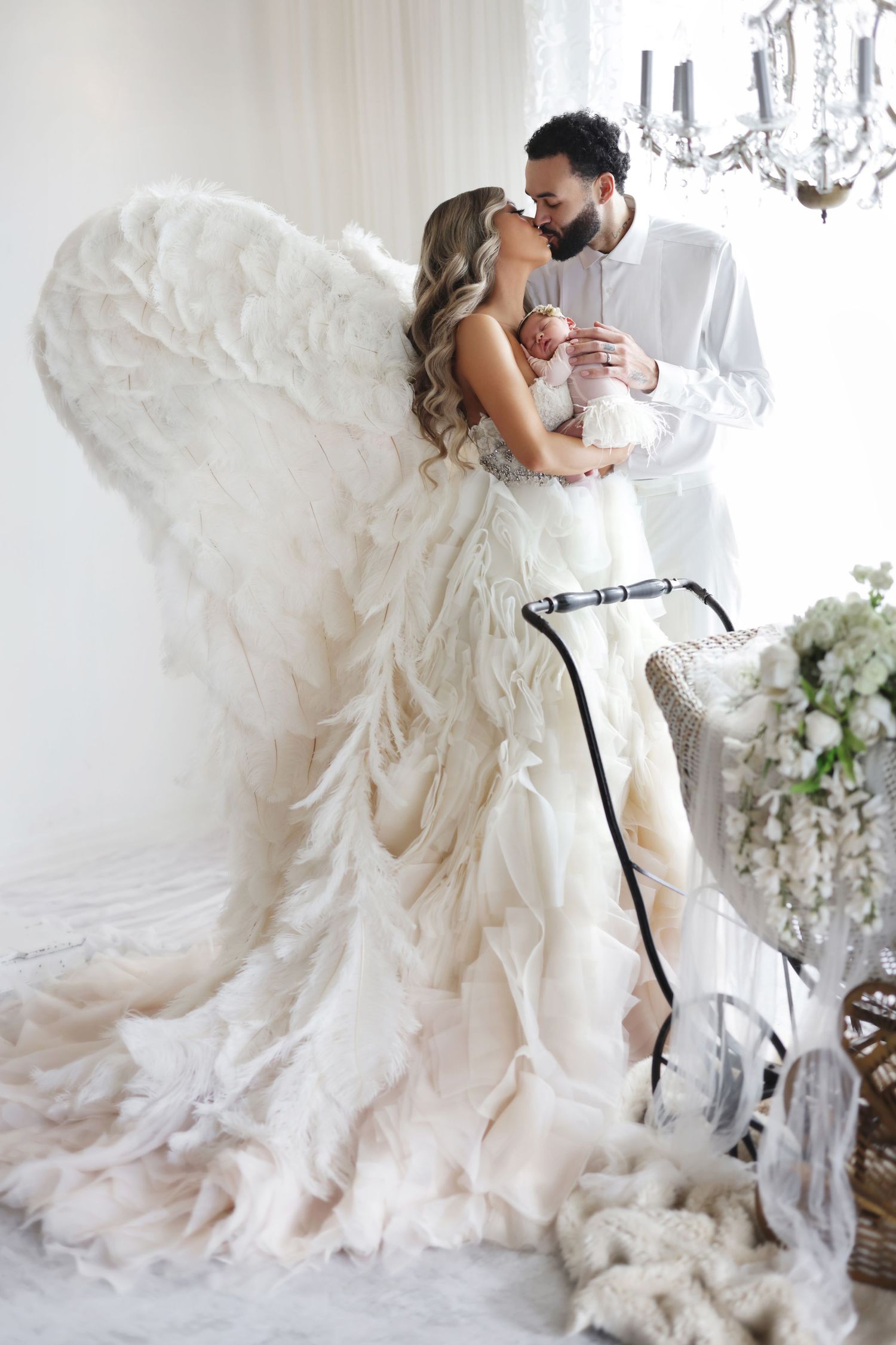 A woman in a feathered angel wing gown holds a newborn while a man in white embraces her in a dreamy white setting.