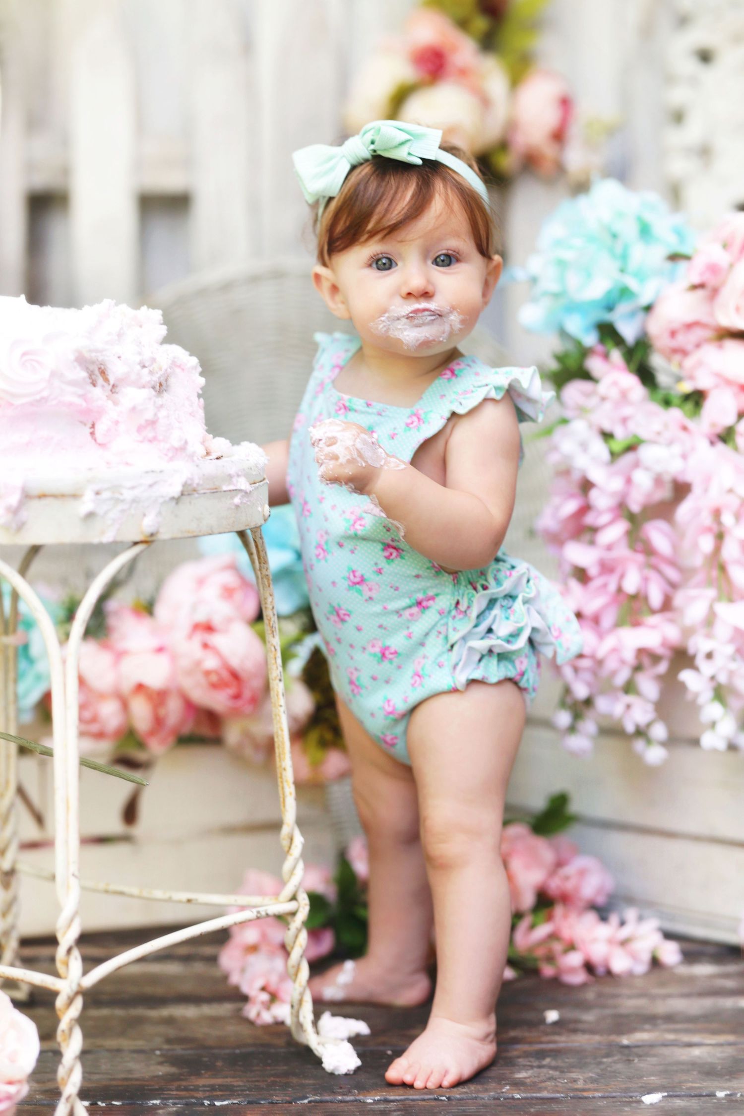 Adorable baby girl with cake on face standing beside smash cake in floral romper.