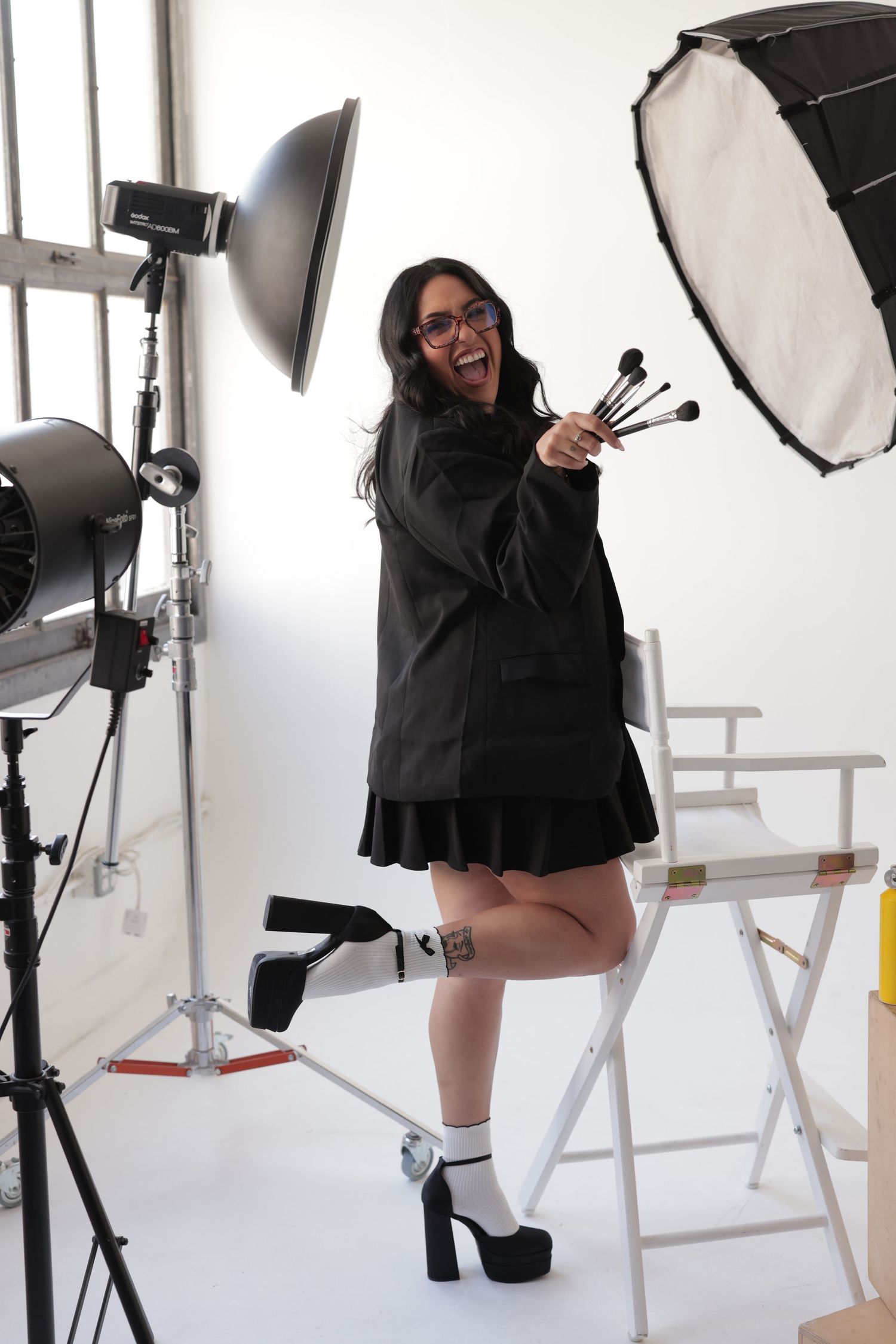Laughing woman in black outfit holding makeup brushes in professional photo studio setting.