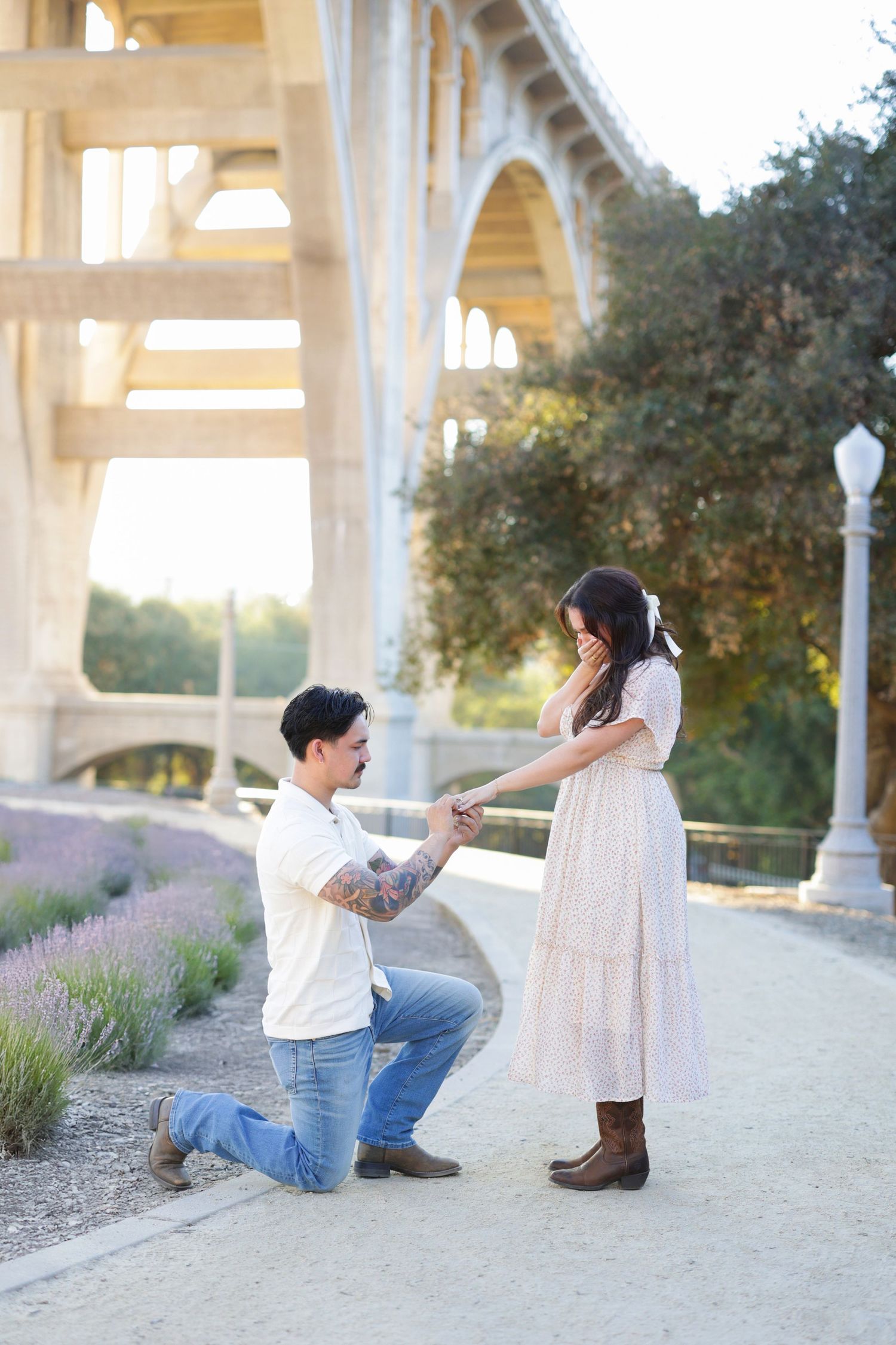 Man kneeling on one knee proposing to emotional woman in pink dress under historic stone bridge outdoors.