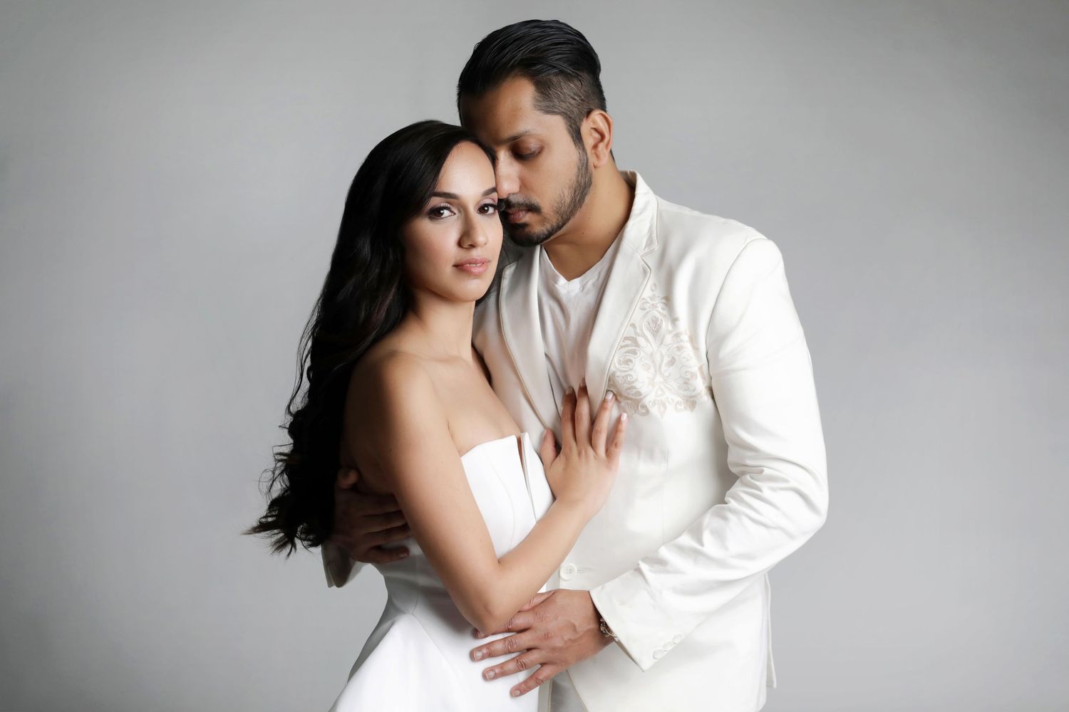 Elegant couple in white outfits posing together, man in white suit embracing woman in strapless white dress.