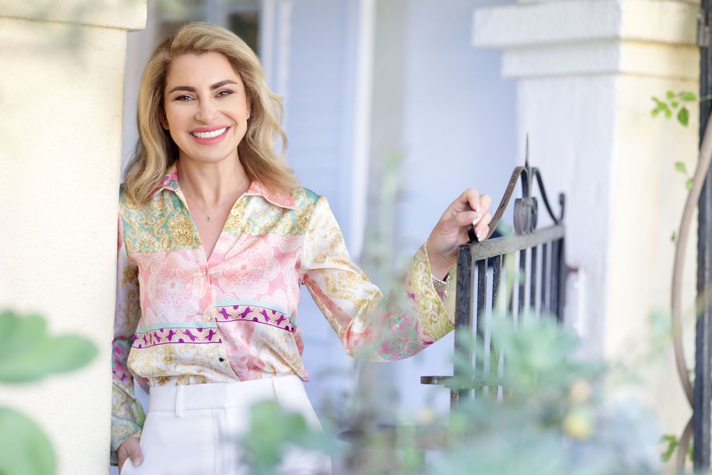 Smiling blonde woman in floral pastel blouse and white pants standing on a sunlit porch.