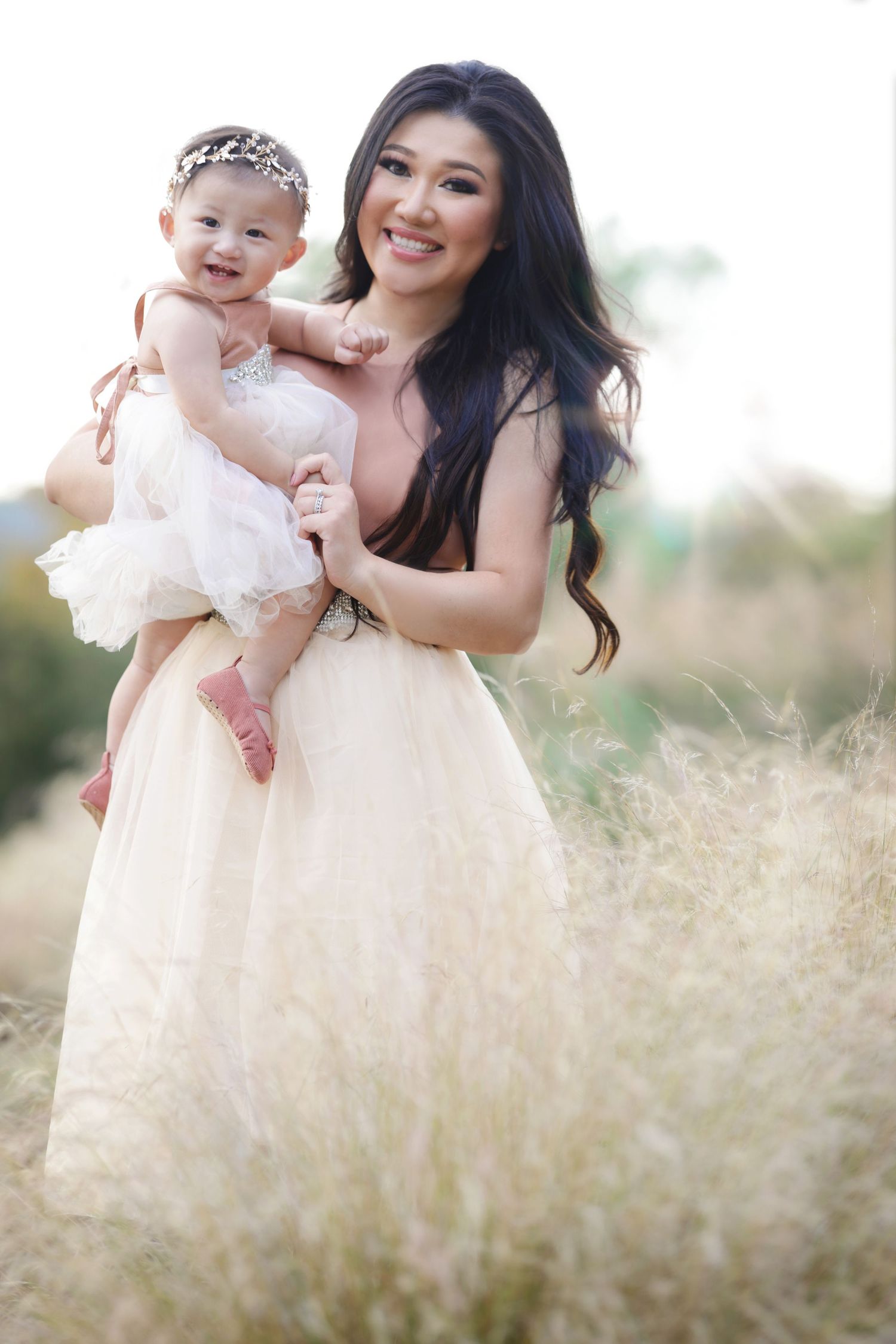 Woman in elegant gown holds smiling baby girl in white dress and tiara outdoors in golden field.