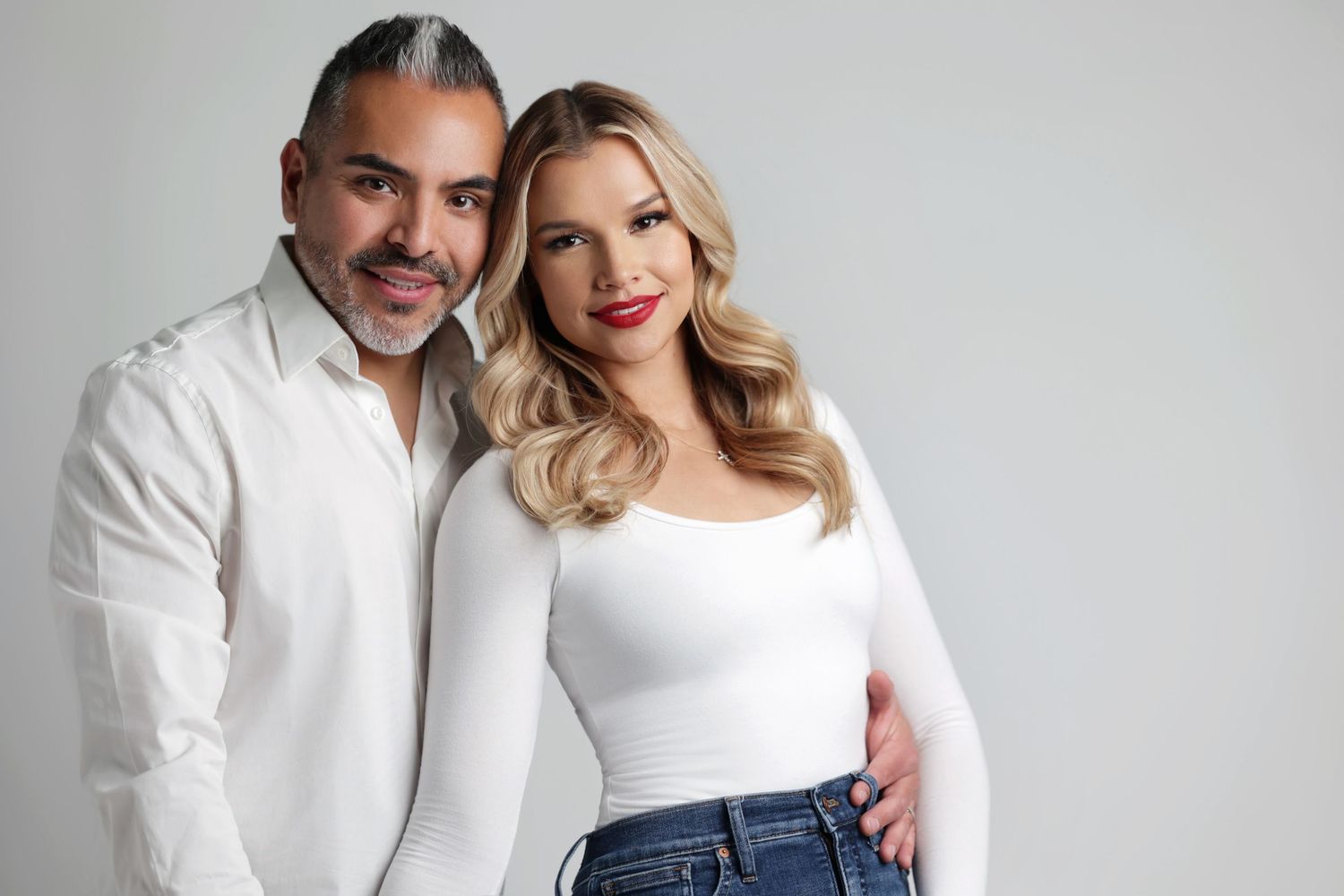 A man and woman in matching white outfits pose together smiling against a light gray background.