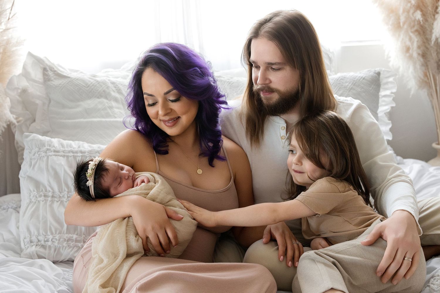Family portrait of parents with purple-haired mom holding newborn baby and toddler daughter sitting together on a bed.