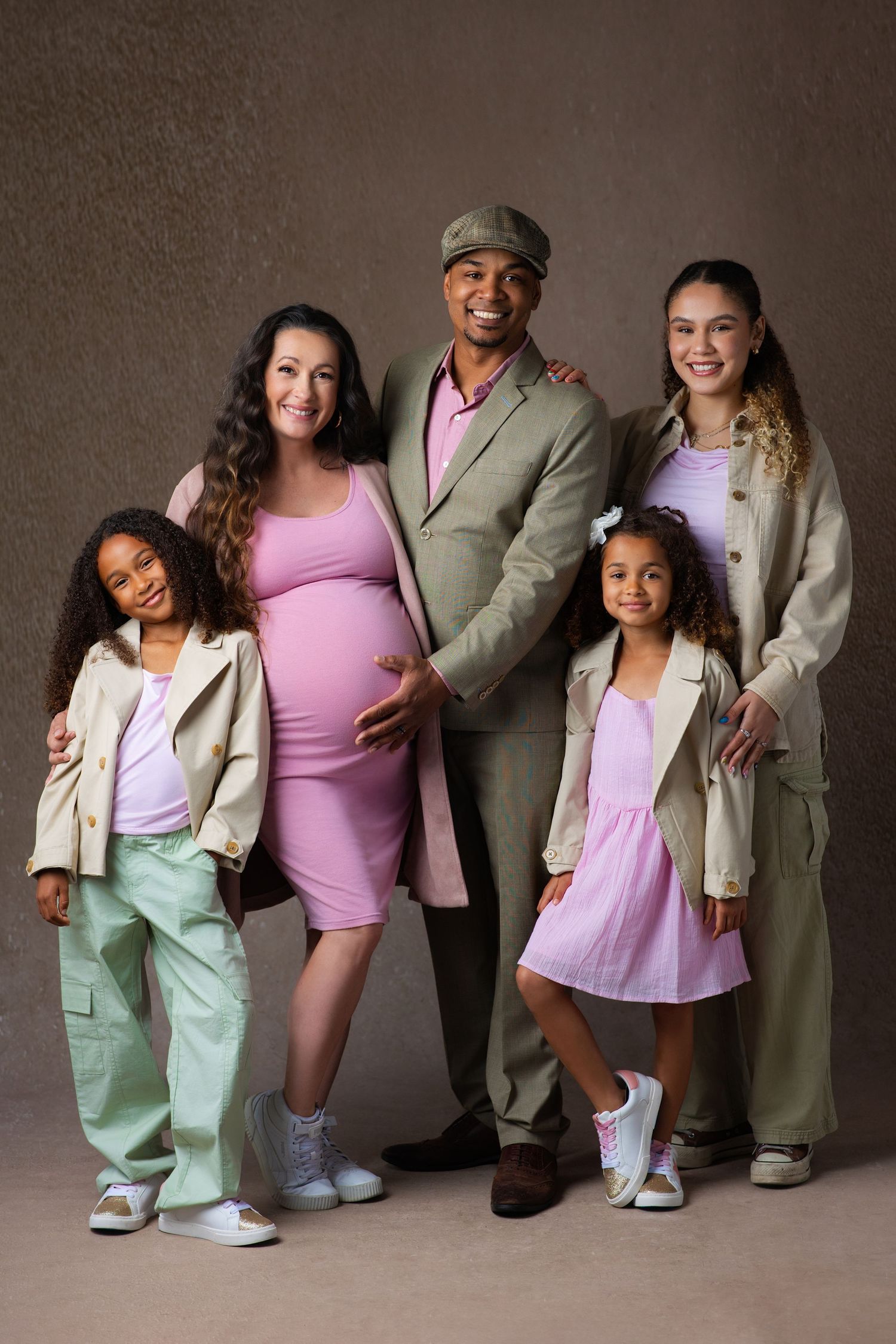 Happy multiracial family with pregnant mother, father, and three daughters posing together in coordinated outfits for a studio portrait.