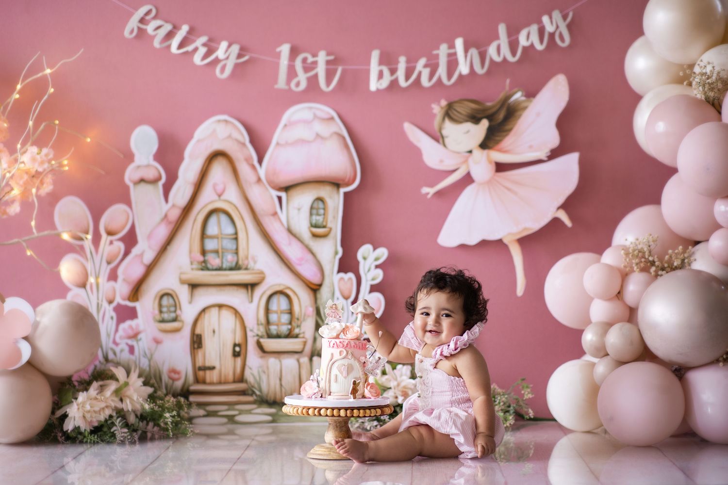 Baby girl celebrating fairy 1st birthday with cake smash, pink balloon arch and fairy tale themed decorations.