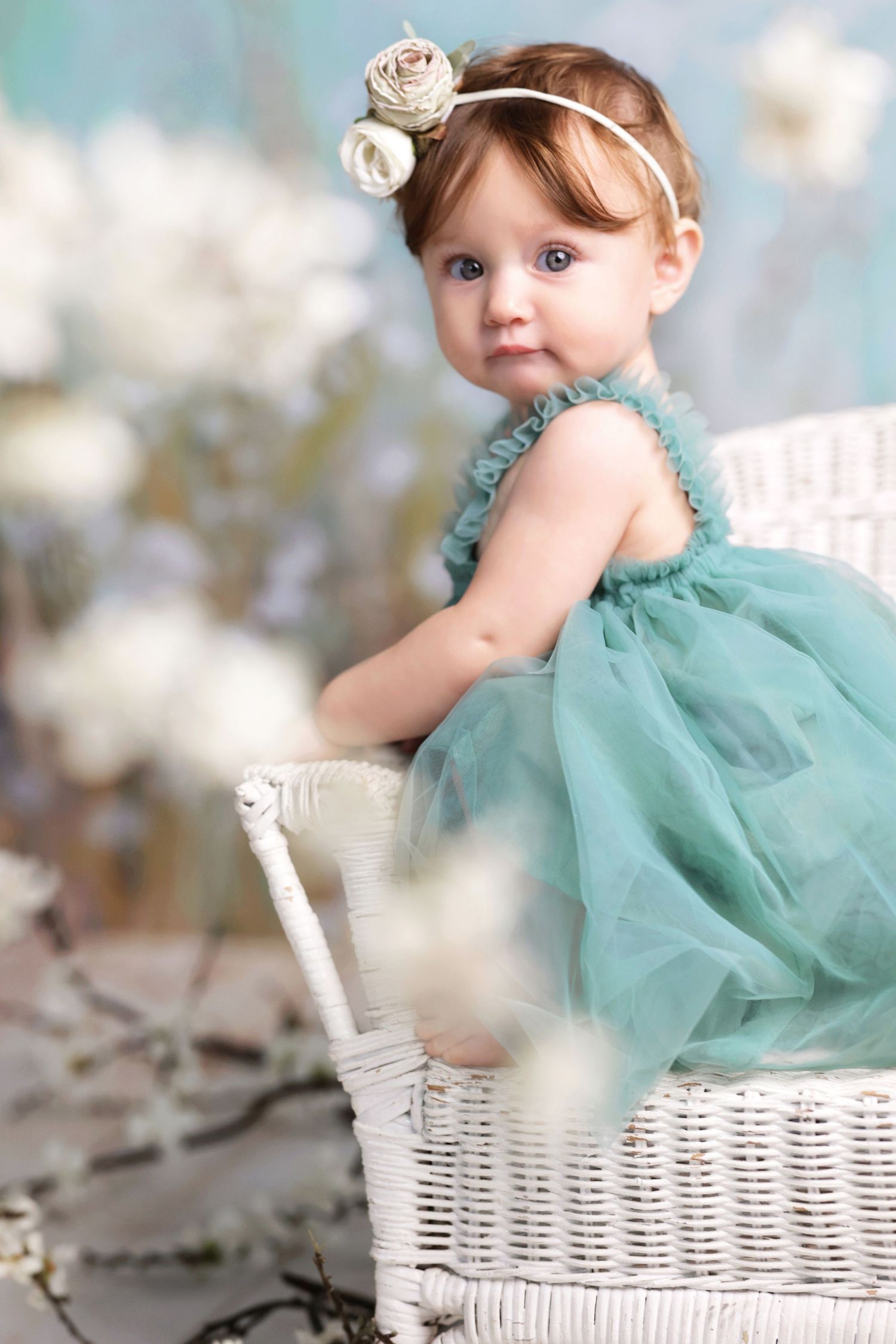 Cute baby girl in teal tulle dress with rose headband sitting in white wicker basket.