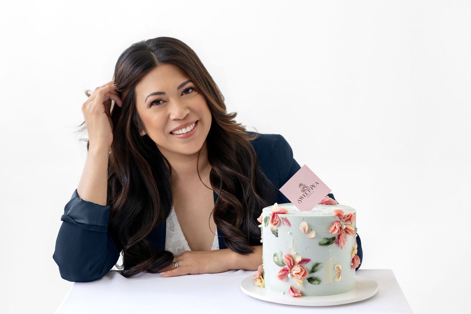 Woman smiling beside a decorated floral two-tier cake on a white background, holding her hair elegantly.