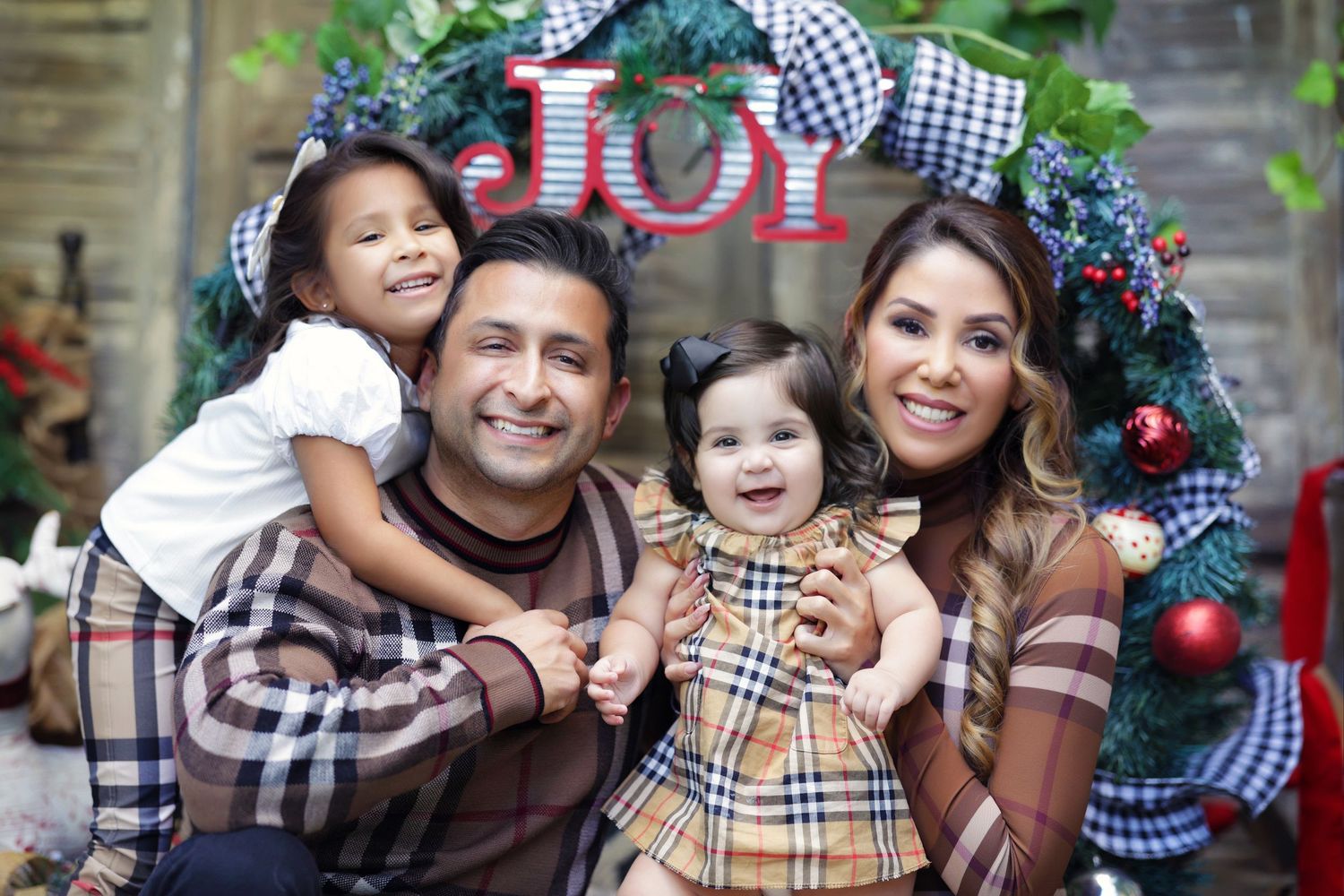Family of four poses joyfully in front of Christmas wreath with JOY sign for festive holiday portrait.