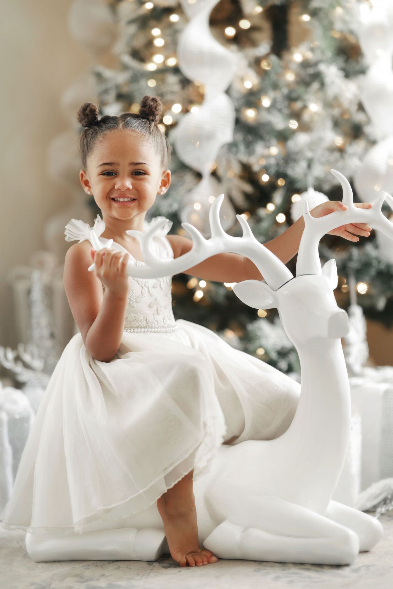 A smiling little girl in a white dress poses with a decorative white reindeer beside a glowing Christmas tree.