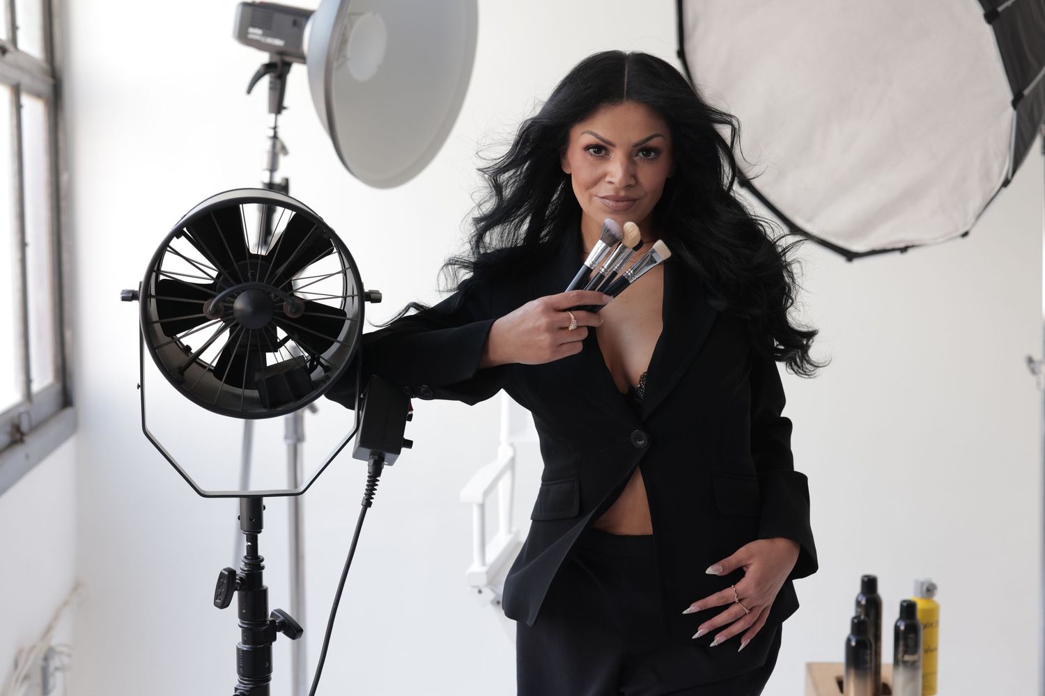 Woman in black blazer posing with makeup brushes in professional photography studio.