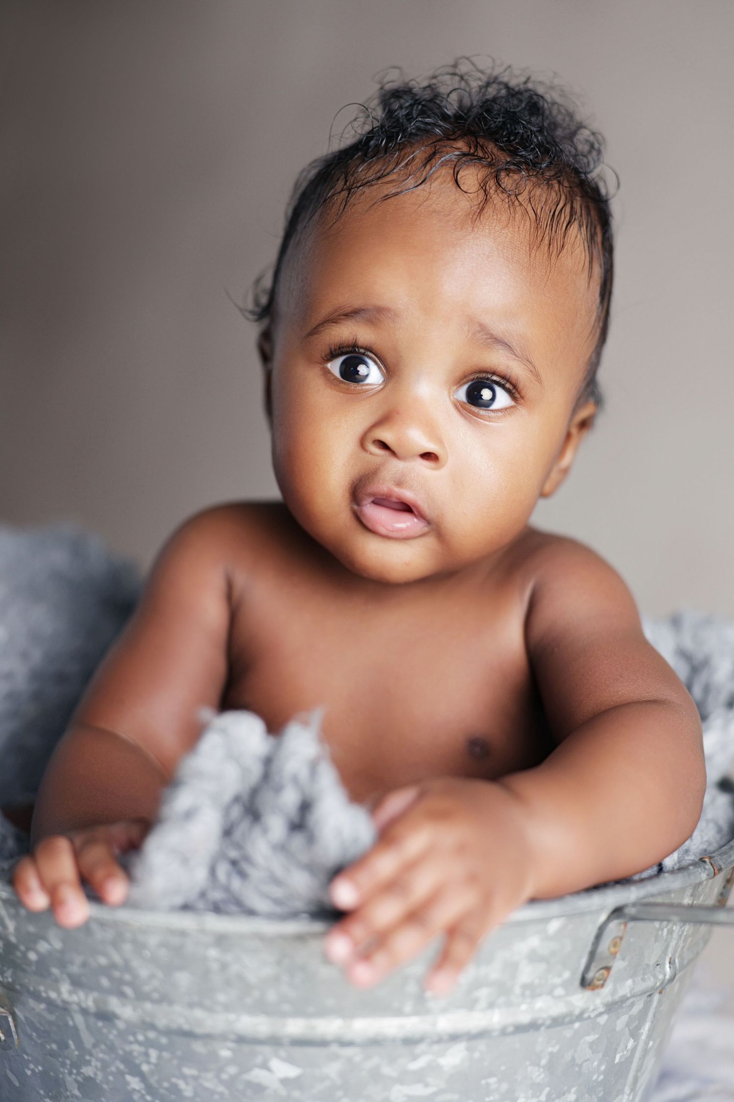 Cute African American baby sitting in a vintage metal tub with a gray blanket, looking up with big brown eyes.