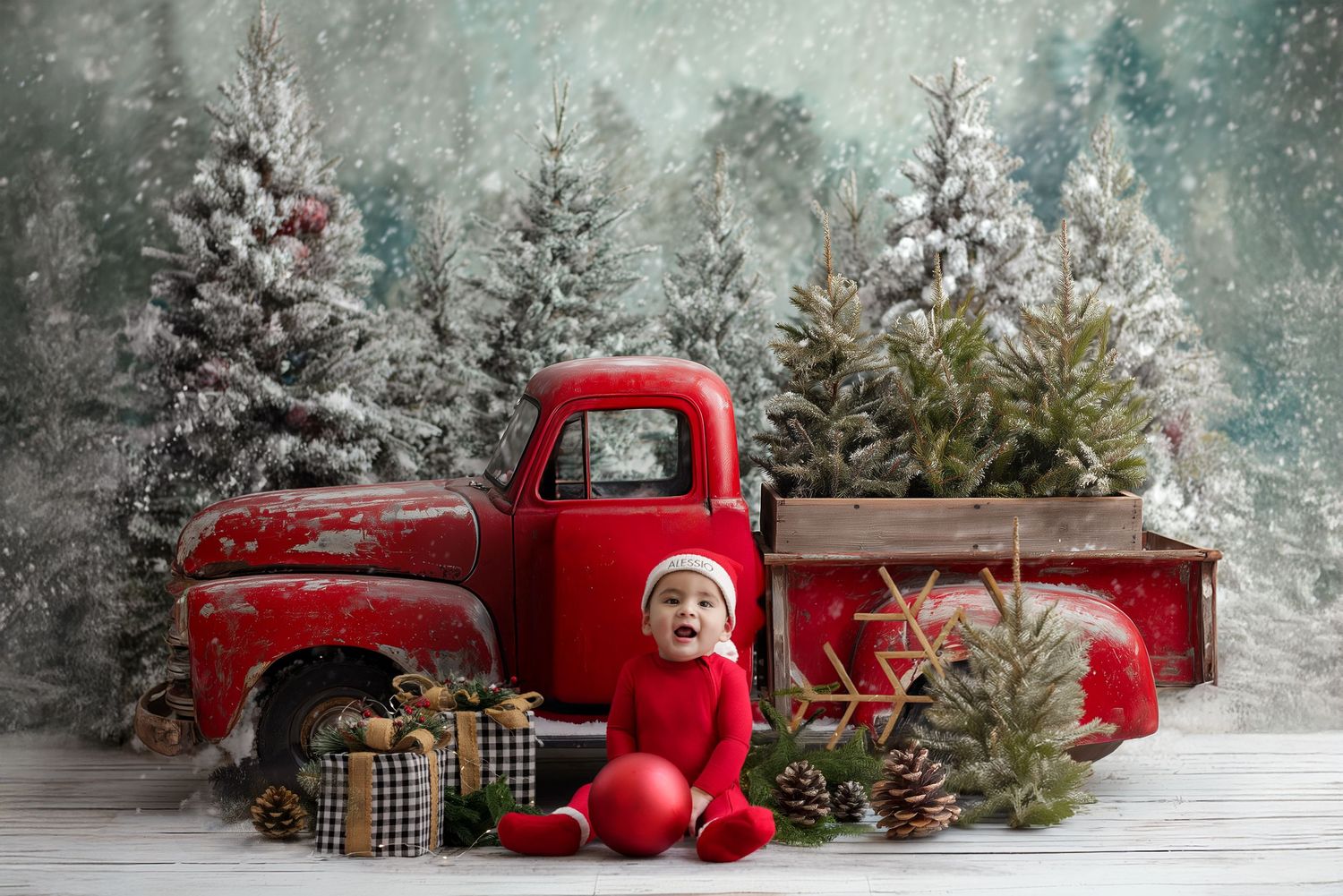 Baby in red outfit sits beside a vintage red truck with Christmas trees and gifts in a snowy holiday scene.