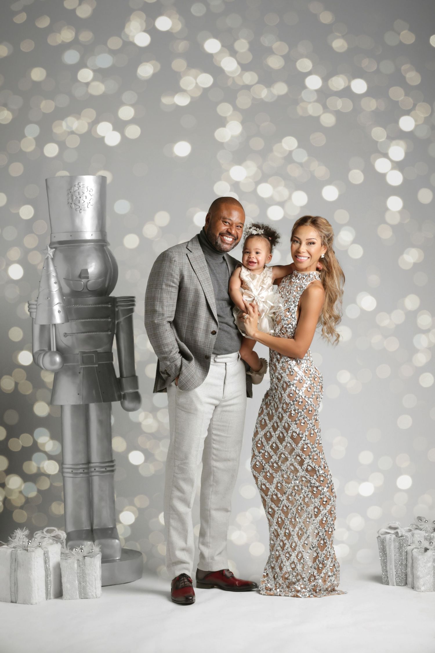 Family of three posing elegantly in silver holiday attire with bokeh lights and nutcracker decor.