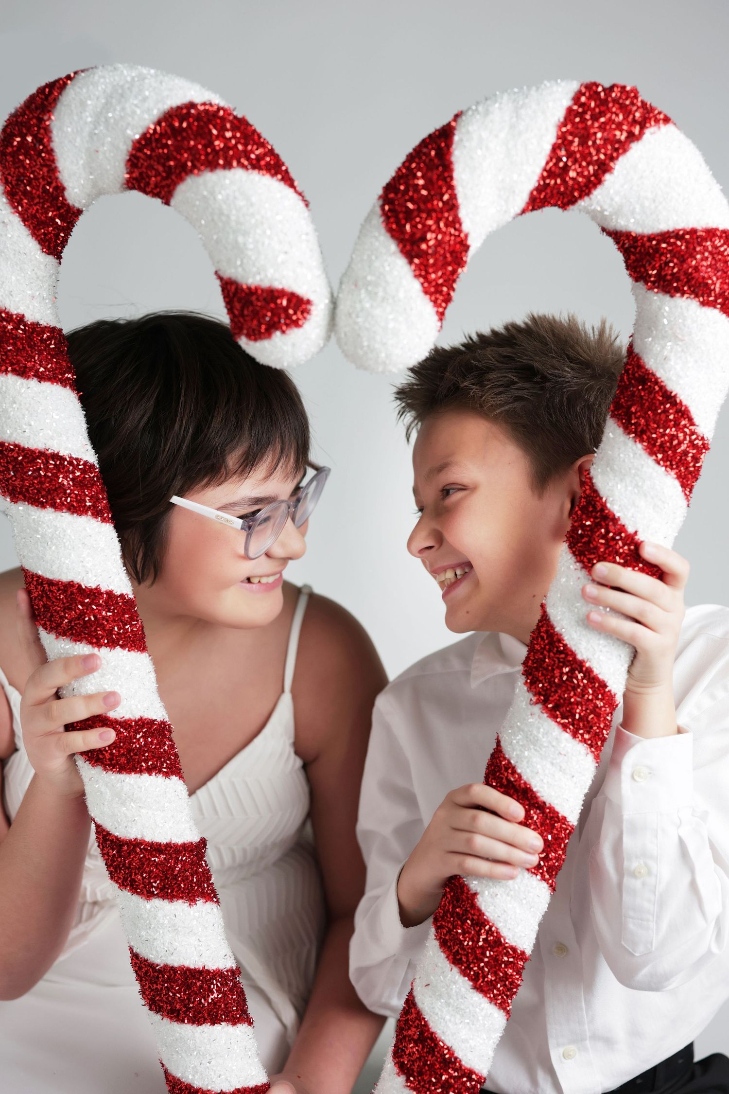 Two children smiling and holding large red and white glittery candy canes in a festive Christmas photo.