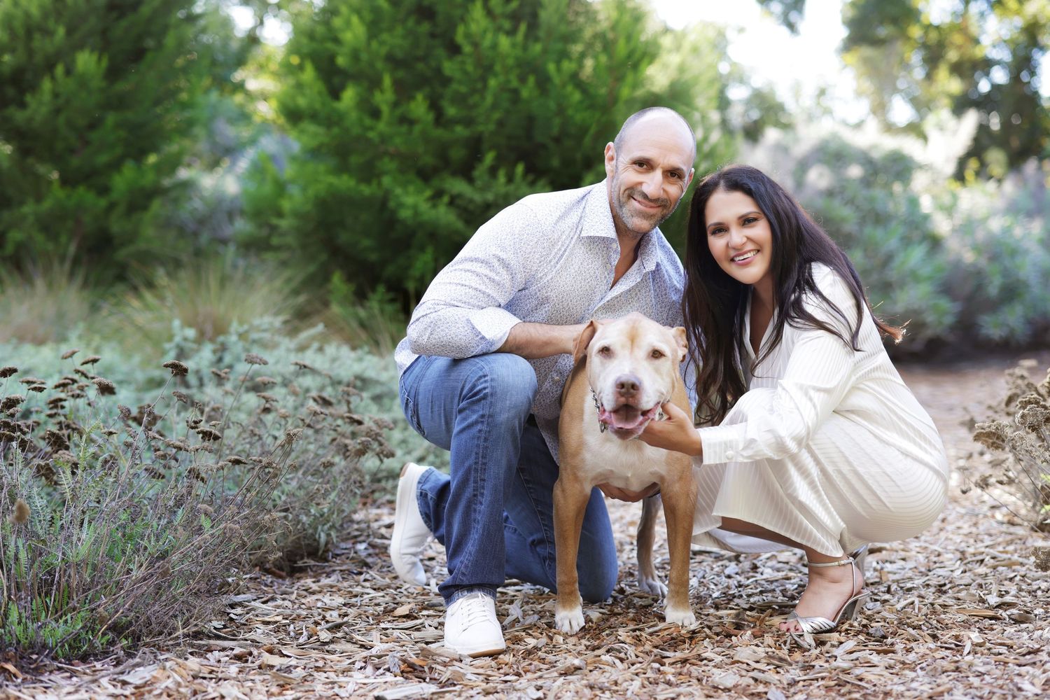 A smiling couple poses outdoors with their tan pit bull dog among lush green shrubs and trees.