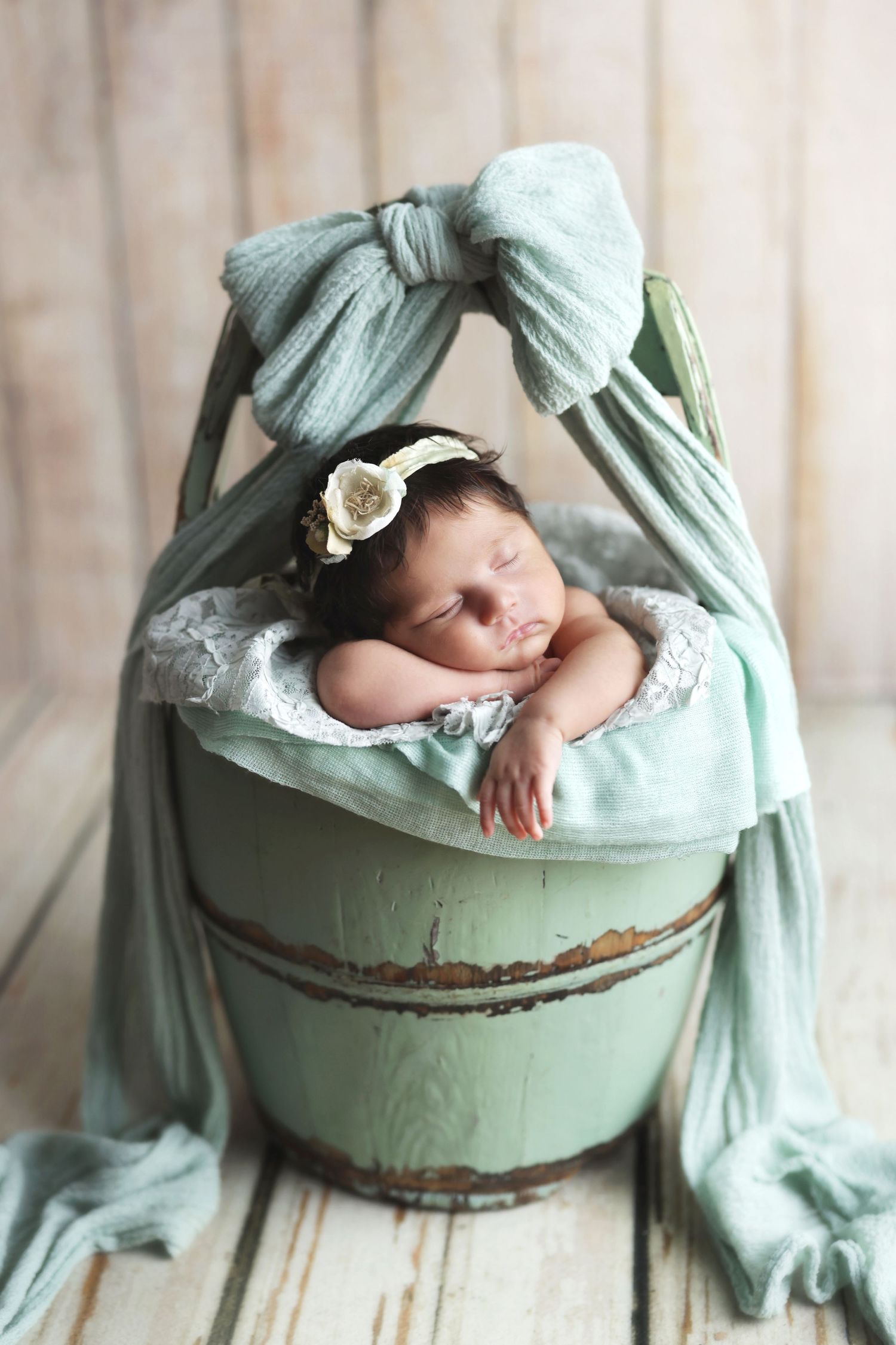 Sleeping newborn baby in a rustic green bucket with teal fabric canopy and floral headband.