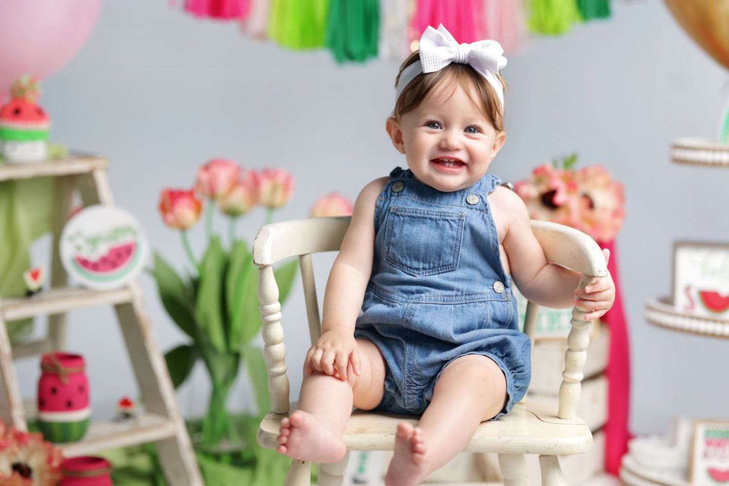 Happy toddler girl in denim overalls with white bow headband sitting in a chair surrounded by watermelon-themed decor.