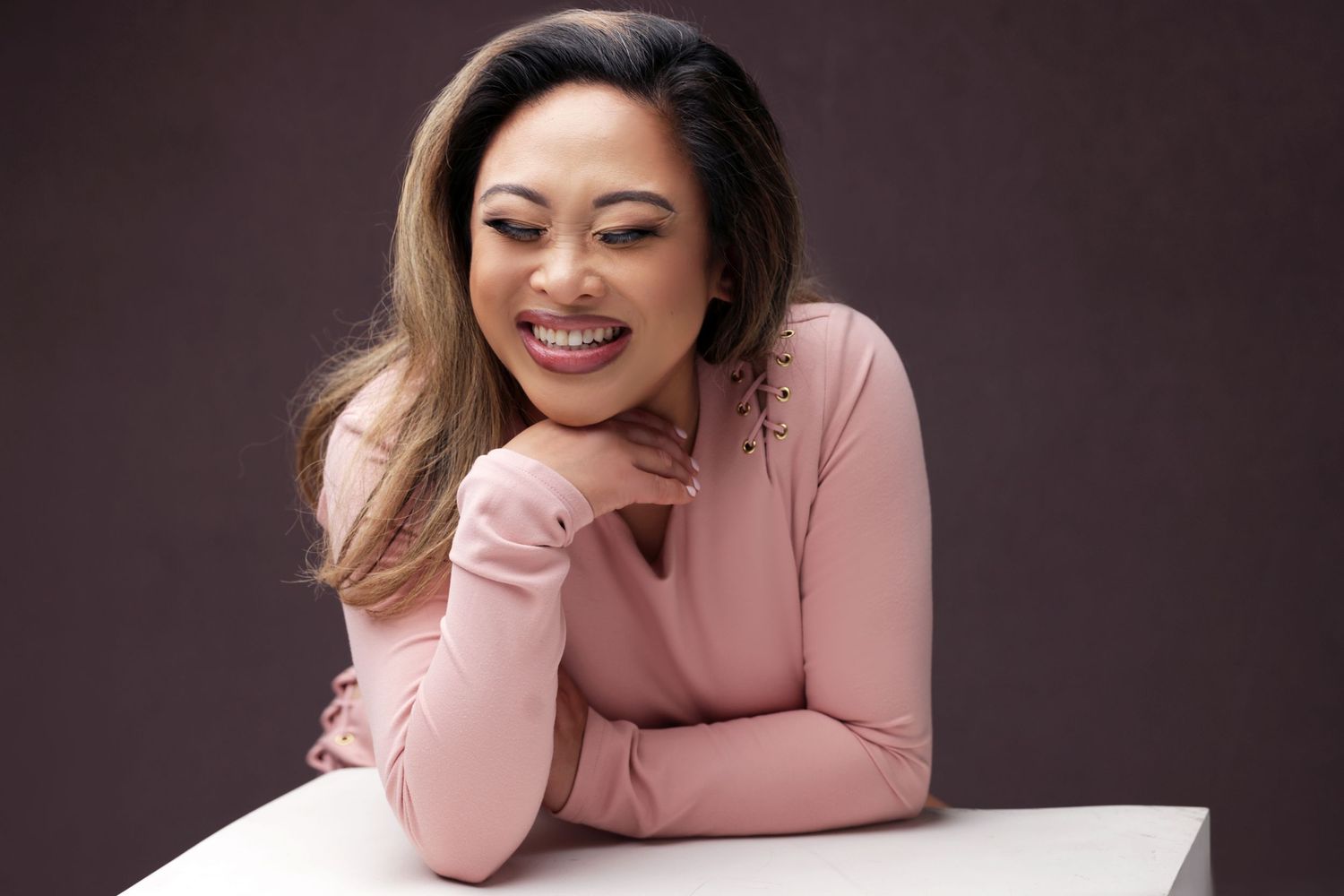 Smiling woman in pink turtleneck top posing elegantly with chin resting on hand against dark background.