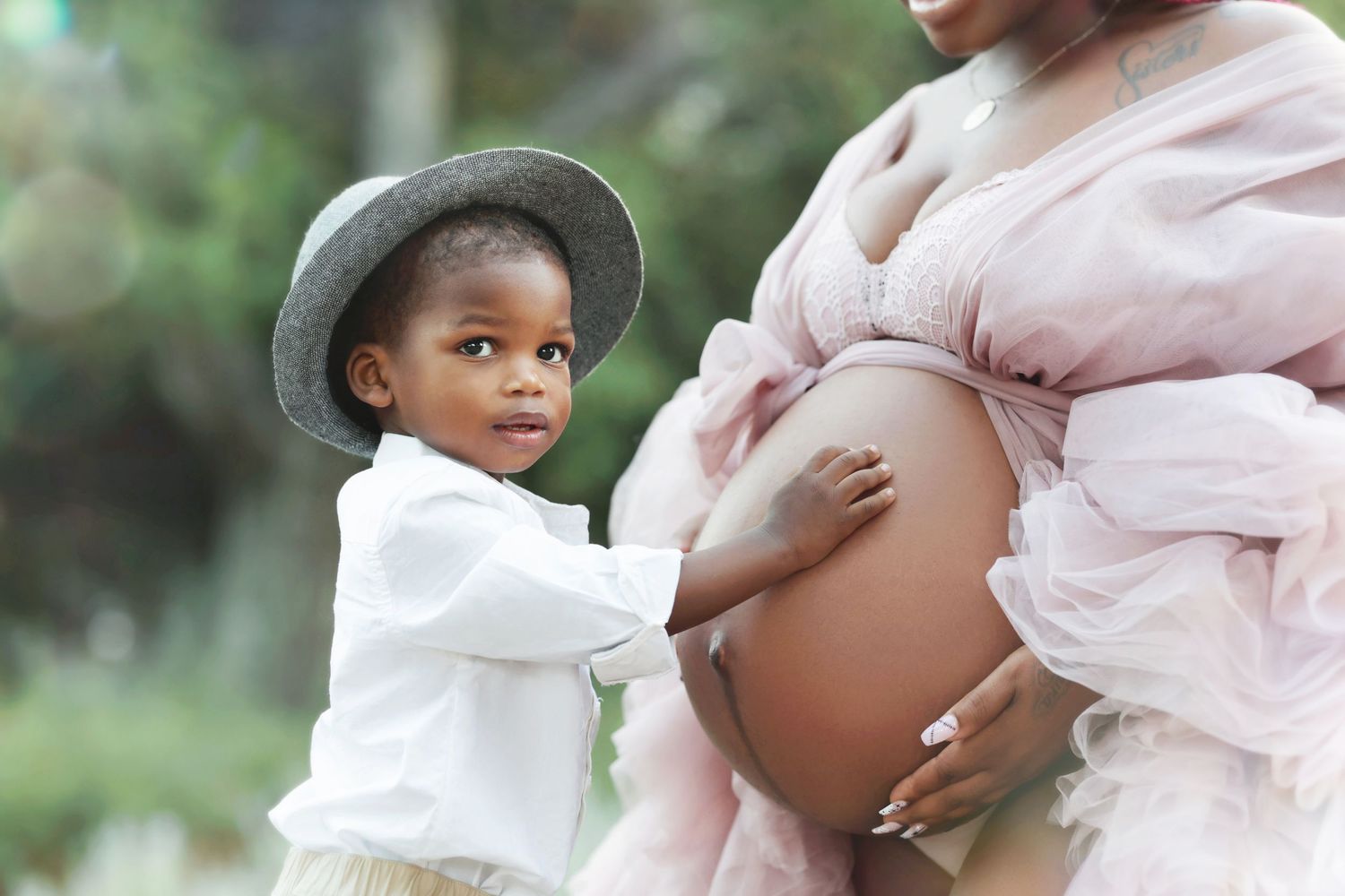 A young toddler boy in a hat touches a pregnant woman's bare belly during an outdoor maternity photo shoot.