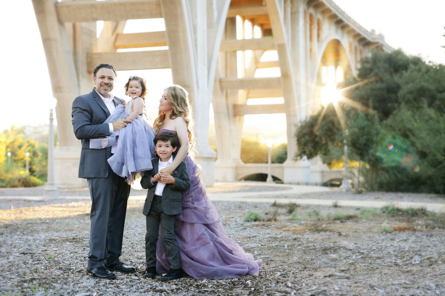 Family of four posing under a bridge at sunset, parents holding children in formal attire on a rocky outdoor setting.