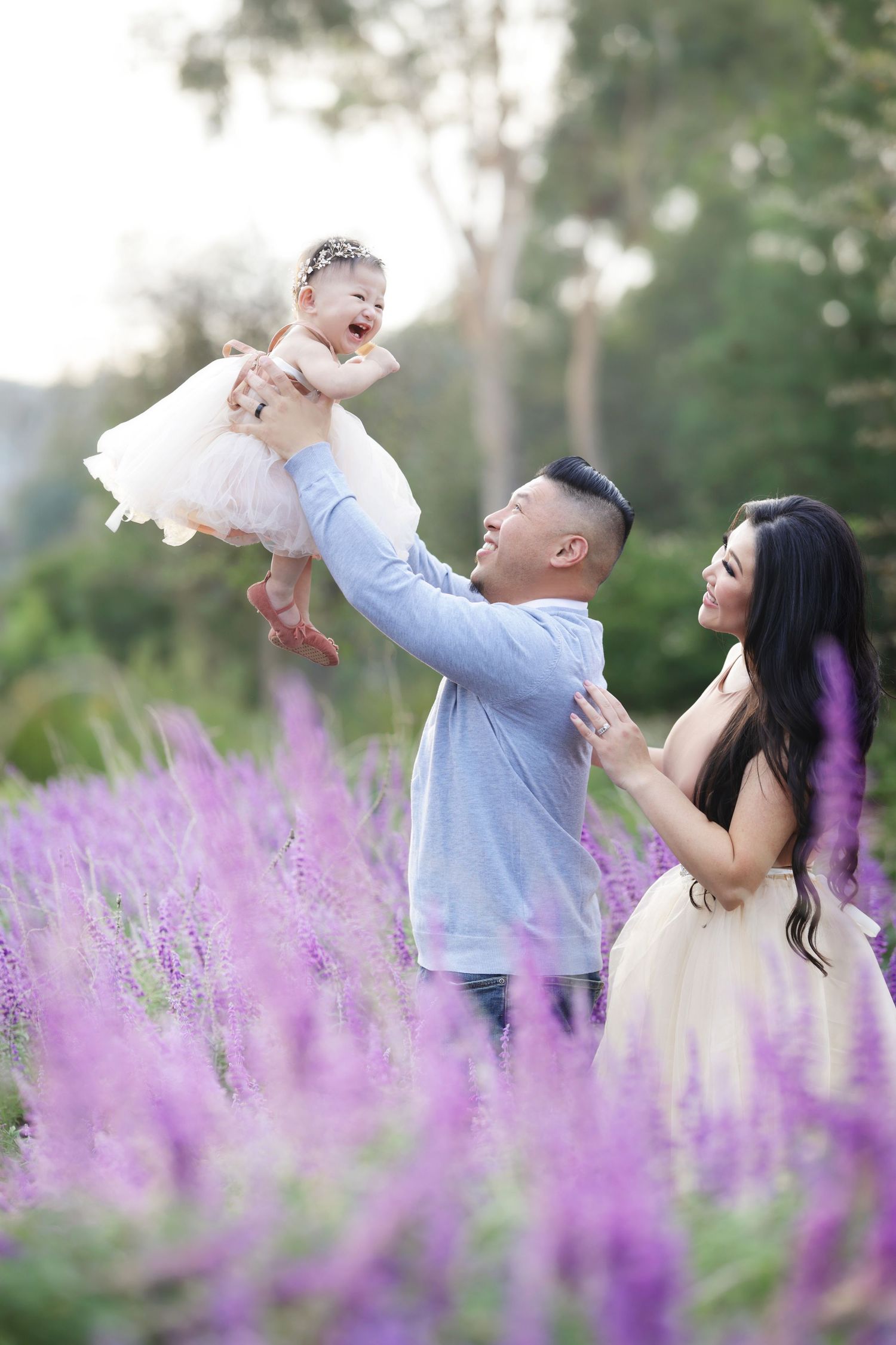 A joyful family poses in a purple flower field, father lifting laughing baby girl while mother watches lovingly.