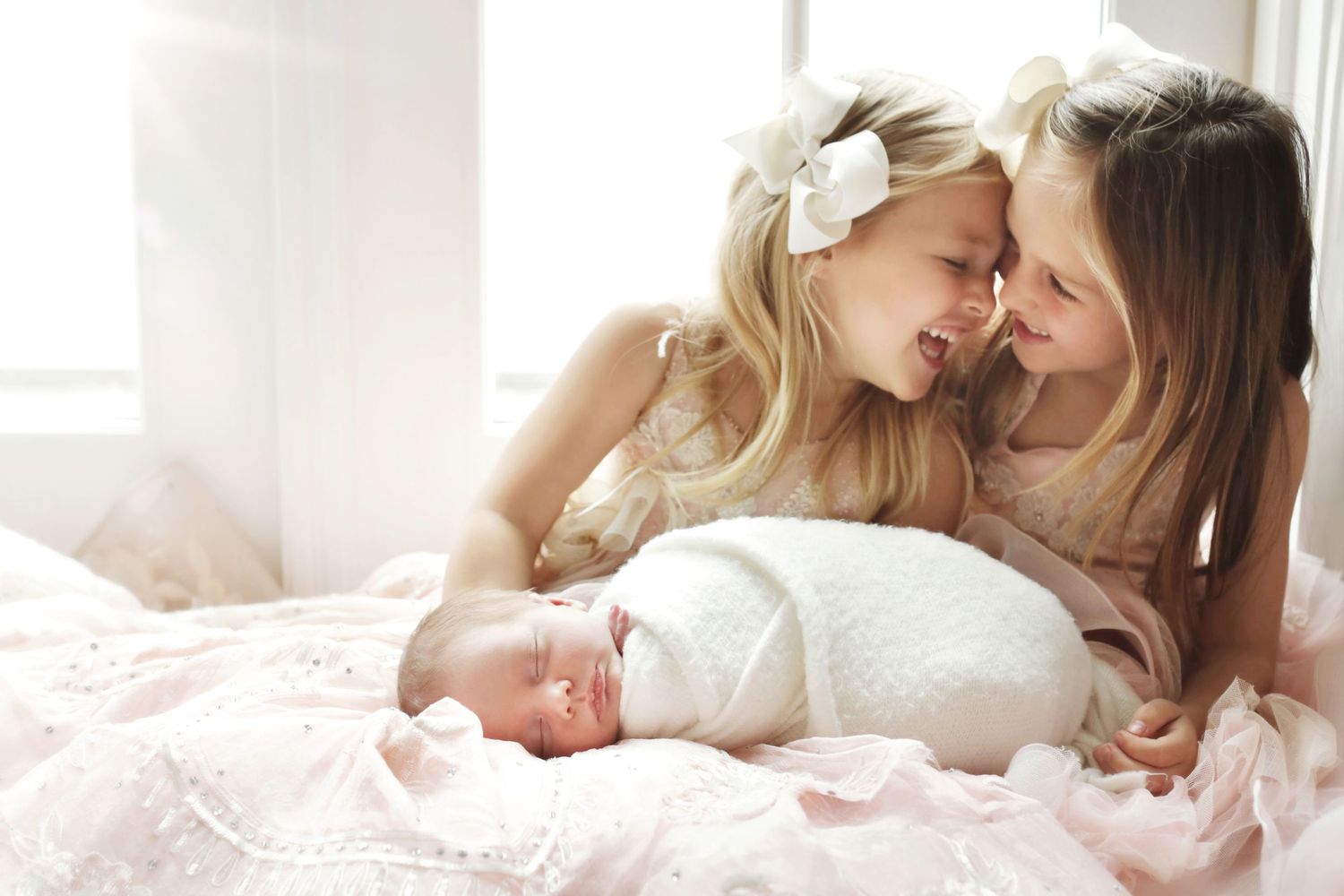 Two young blonde sisters in pink dresses tenderly nuzzle foreheads over their swaddled newborn sibling on a soft bed.