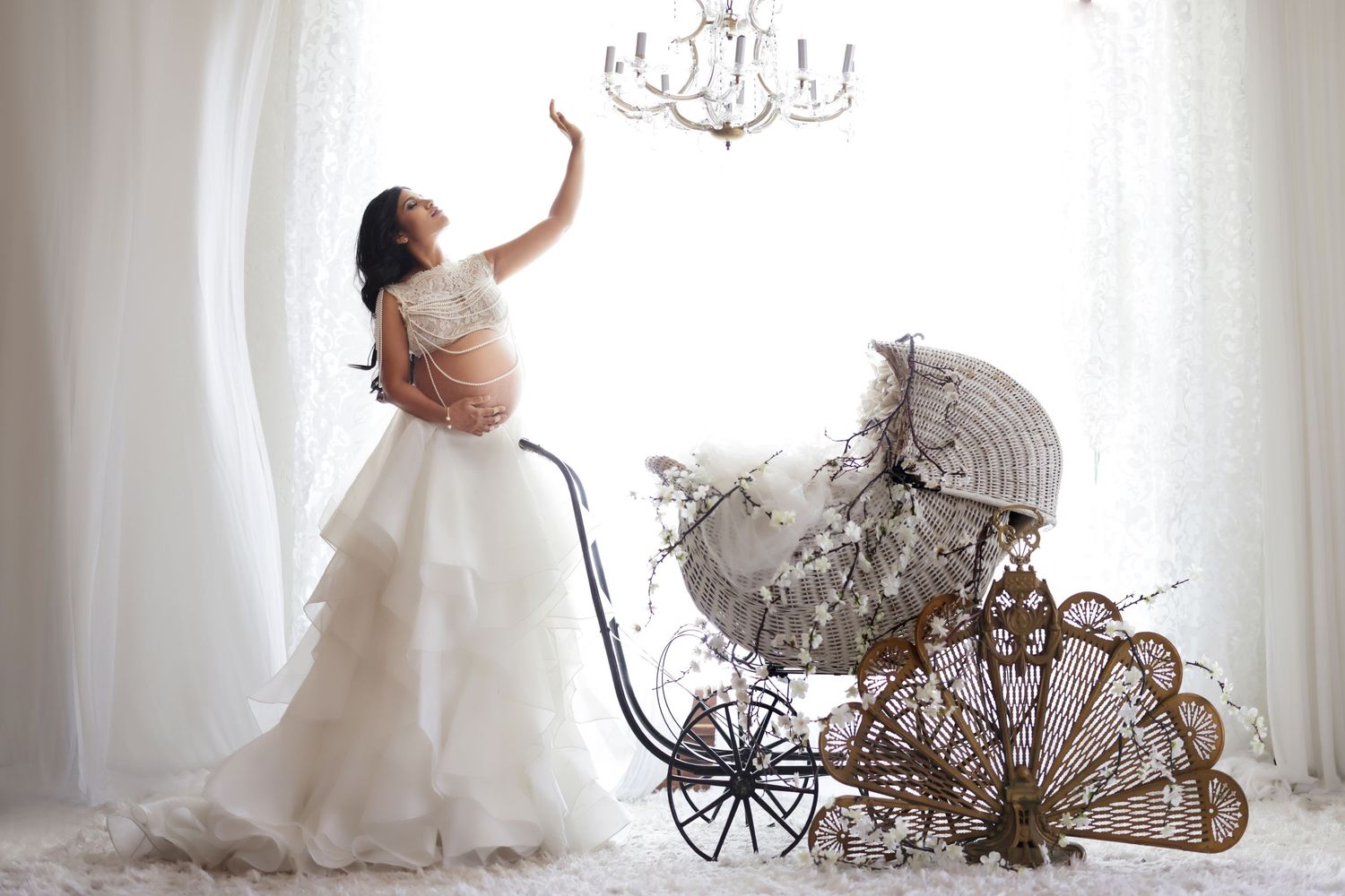 Pregnant woman in white ruffled gown reaching toward chandelier beside vintage ornate baby carriage in bright studio.