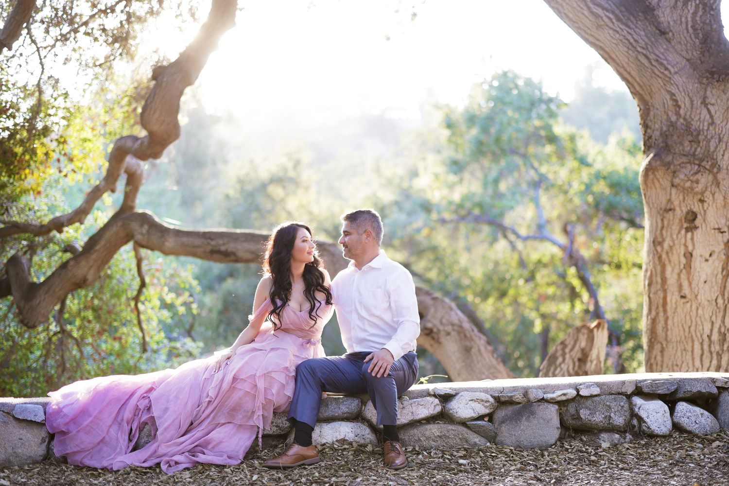 Couple sitting on a stone wall under a tree, woman in a flowing lavender gown, man in white shirt, surrounded by lush greenery.