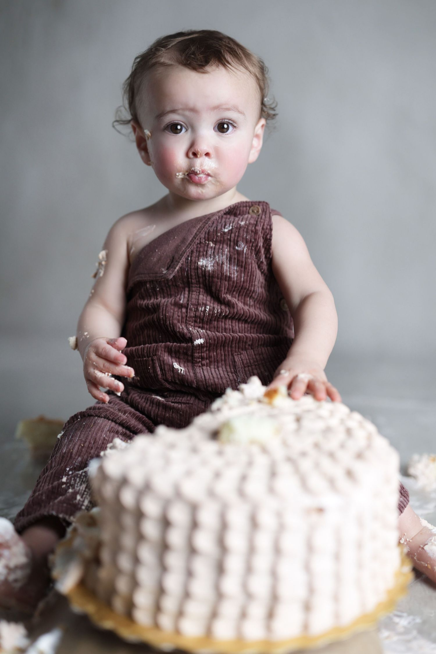Adorable baby in brown sparkly romper smashing first birthday cake, with frosting on face.
