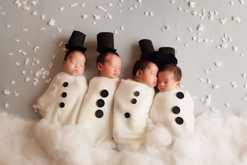 Four newborn babies dressed as snowmen in white swaddles with black hats and buttons, sleeping peacefully together.