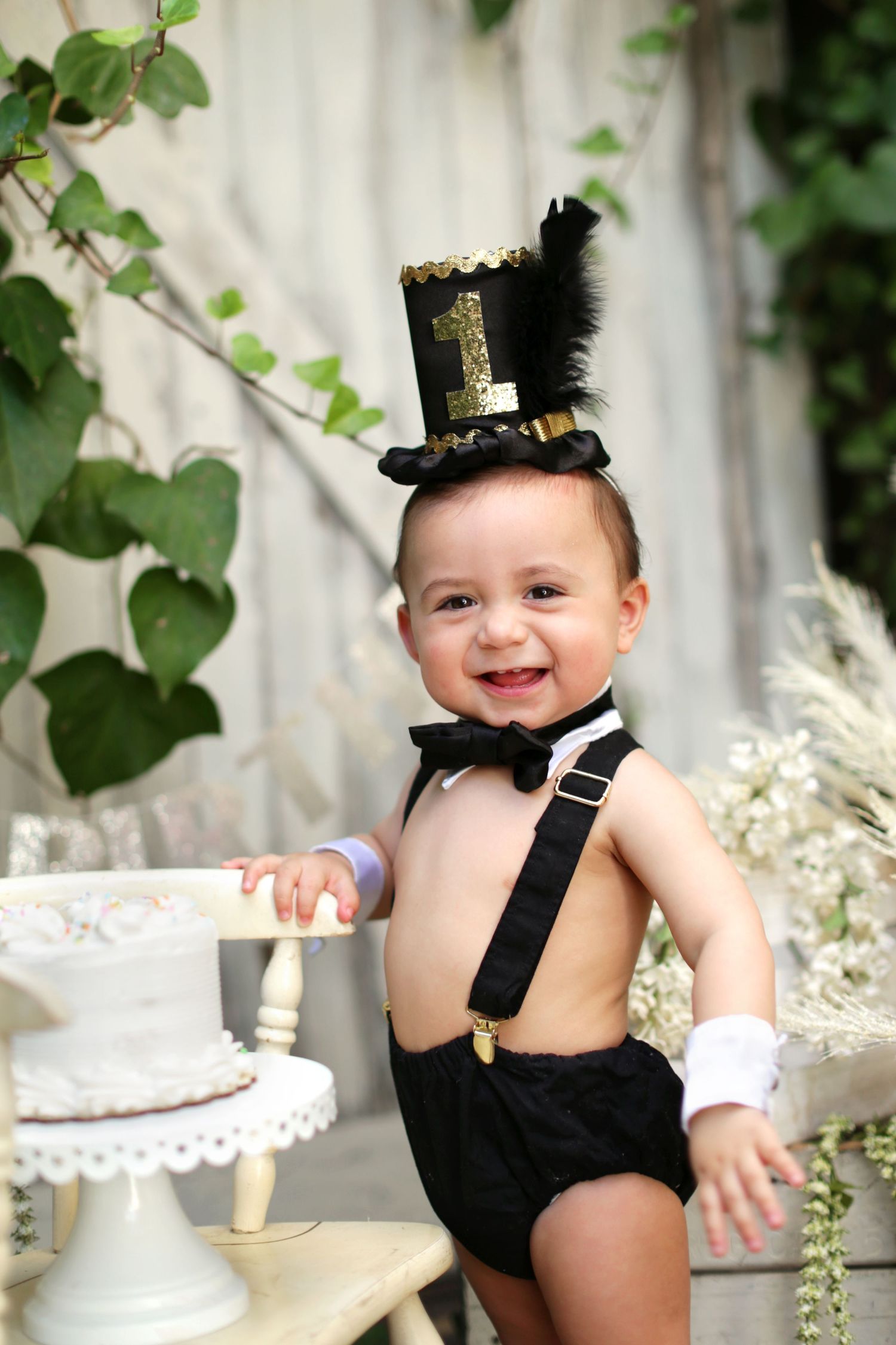Adorable baby boy in tuxedo suspenders and top hat poses beside white cake for first birthday.