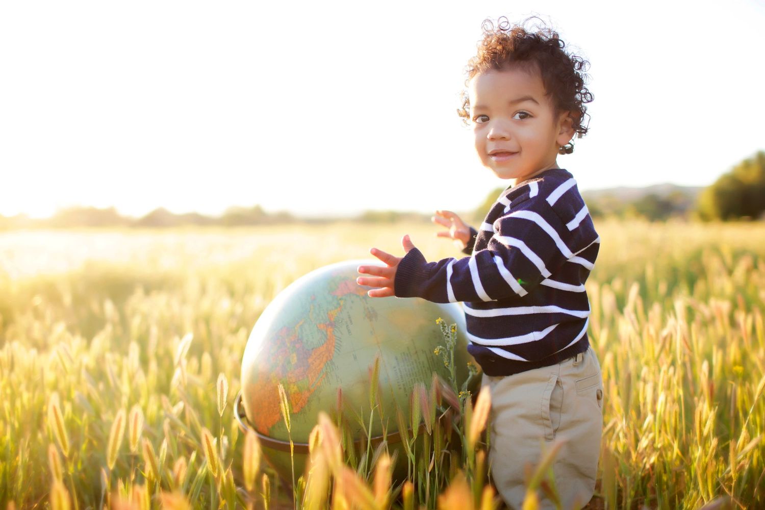 Young child holding a large globe in a sunlit golden field, smiling joyfully at the camera.